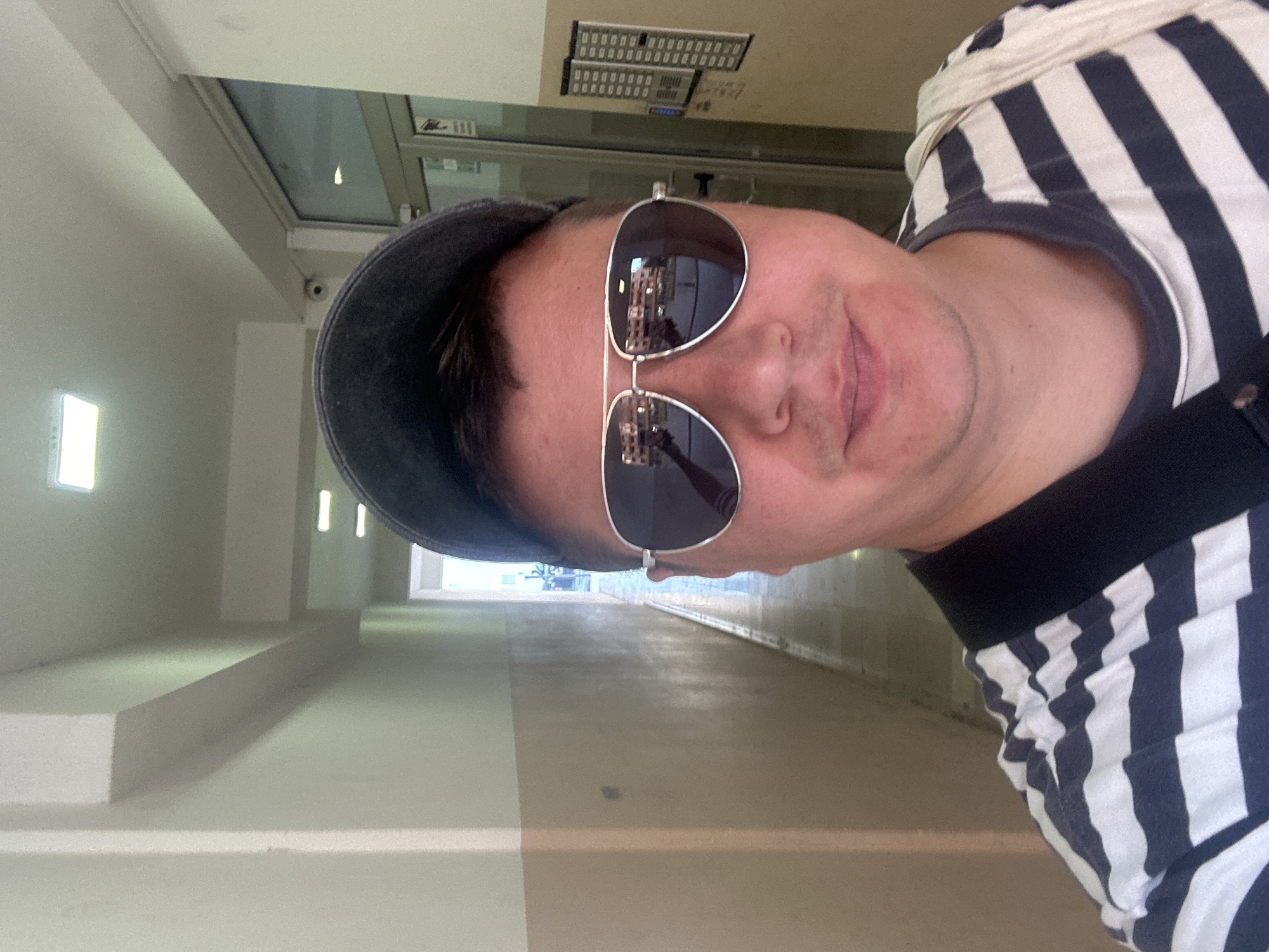 The image is a selfie taken in an indoor hallway, likely in Budva, Montenegro. A person wearing a dark baseball cap, aviator sunglasses, and a navy and white striped t-shirt is prominently featured. The hallway has beige and white painted walls and fluorescent ceiling lights. The floor appears to be tiled, with a long, narrow corridor stretching into the background. On the right wall, a door with glass panels is visible, and next to it, a building's intercom system with multiple buttons is mounted. The reflection in the sunglasses shows a building with balconies, suggesting the hallway might be part of a residential building or a hotel in a populated area. A light-colored bag is slung over the person's shoulder.