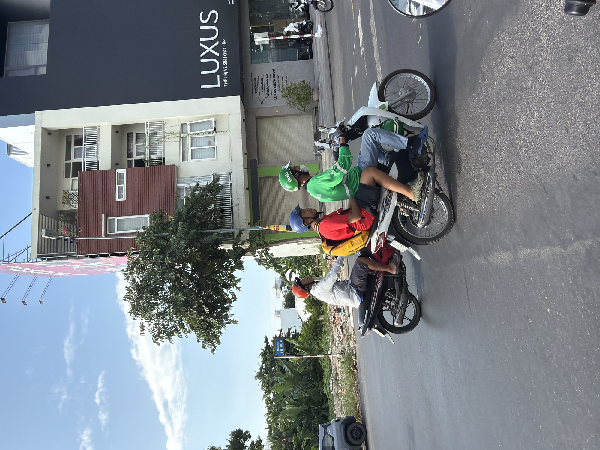 Two men are riding on a white motorcycle on a street in Nha Trang, Vietnam. The man in the front is wearing a green Grab uniform, a blue helmet, and a face mask. He is operating the motorcycle. The man behind him is wearing a red shirt, a blue helmet, and a yellow backpack. He is looking to the left. A black car is parked on the left side of the street. There is a street sign with Vietnamese text on it. In the background, there is a building with multiple floors and advertisements. The sky is blue with some clouds.