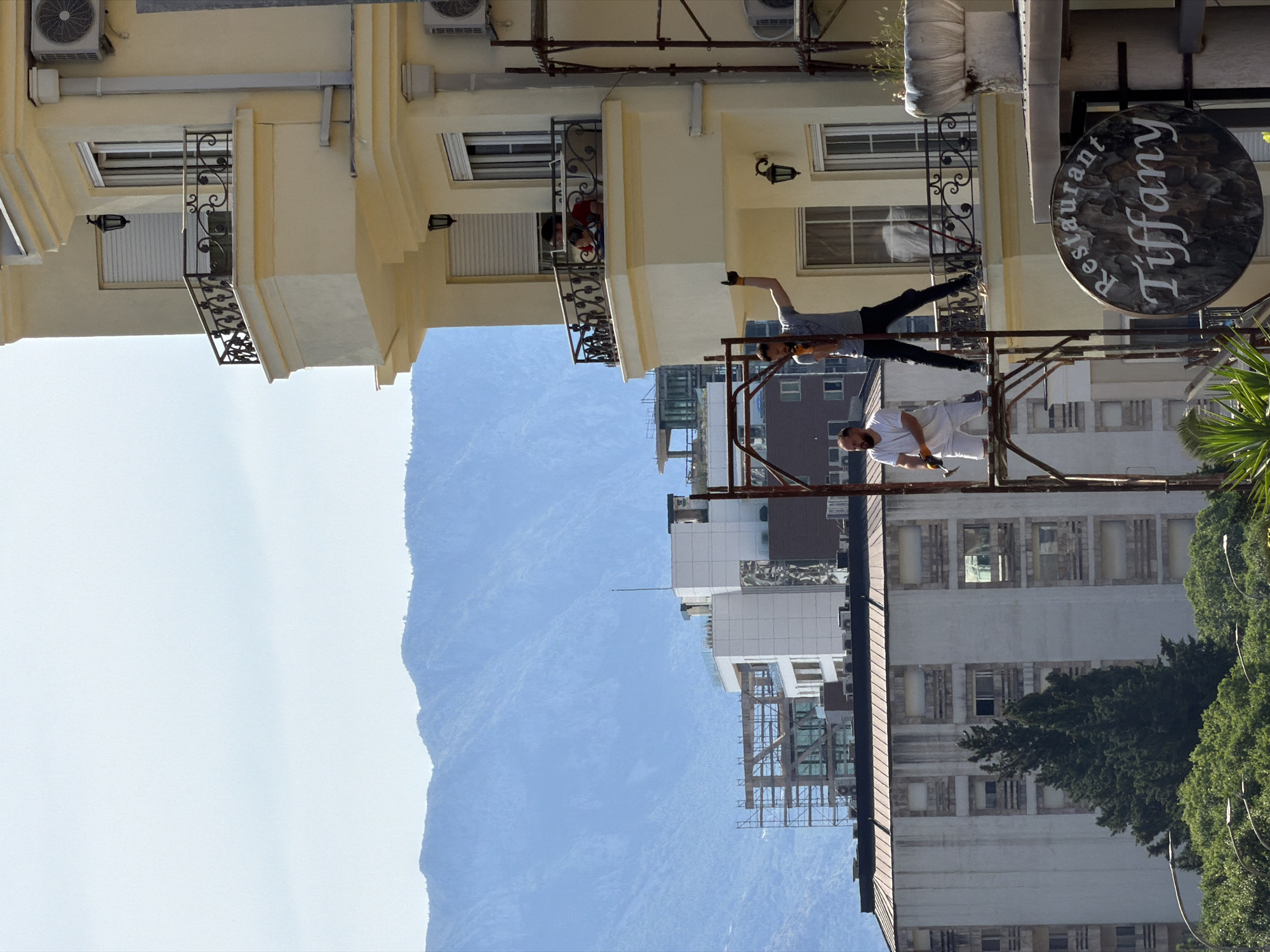 The scene depicts an outdoor setting in Budva, Montenegro. Two men stand on scaffolding against a backdrop of buildings and mountains. One man is dressed in white, seemingly working with tools, while the other is wearing grey and black. The "Tiffany Restaurant" sign is visible. A person is also visible on a building's balcony.