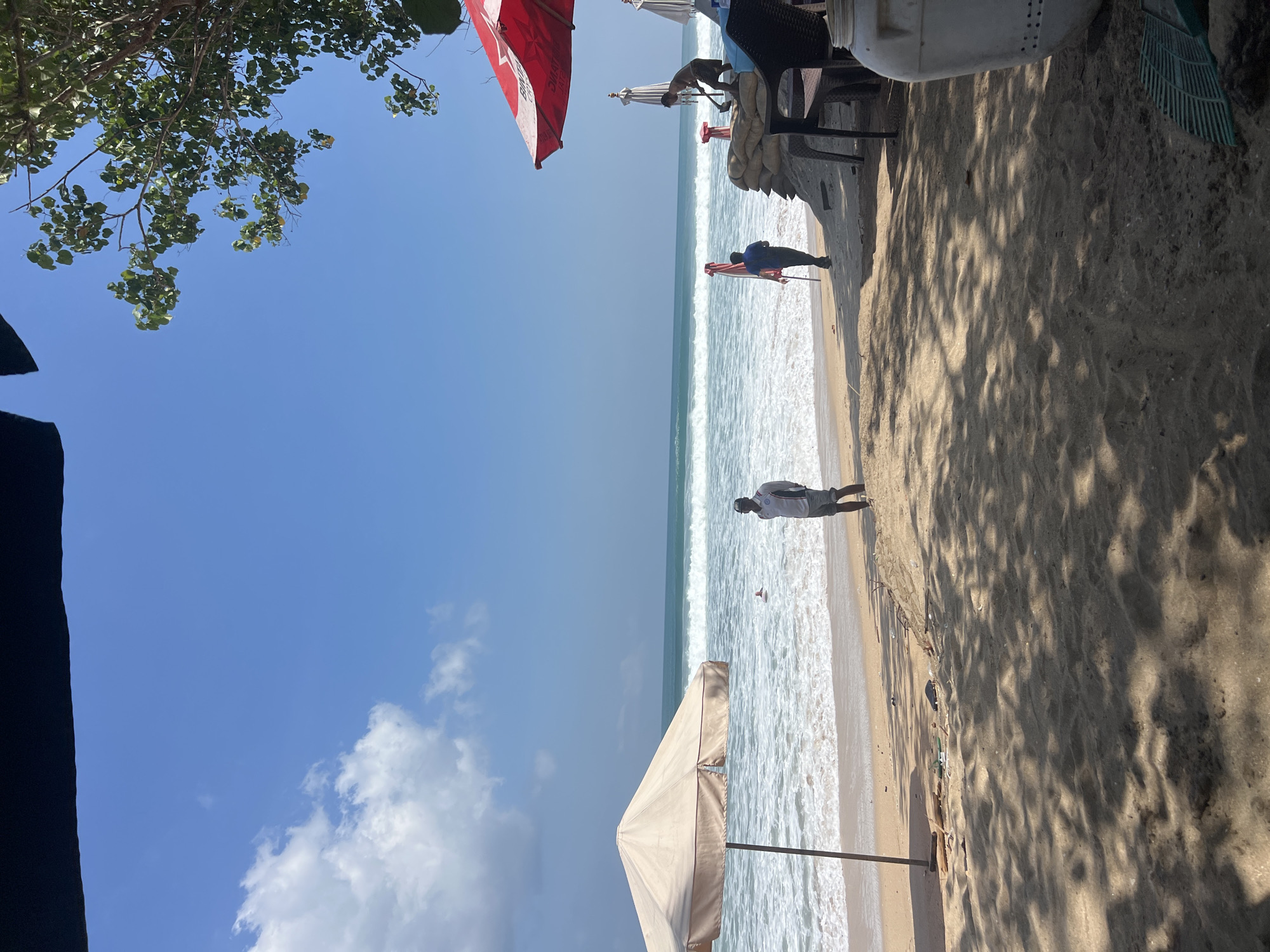 A wide shot captures a beach scene in Kuta, Indonesia on a clear, sunny day. The foreground is dominated by sandy beach and scattered debris. In the middle ground, the ocean waves crash onto the shore, with a single person visible in the water. Two men are on the beach; one is standing by a large umbrella and the other appears to be working with sandbags. A red and white umbrella stands to the right, shading a seating area with chairs. The sky is bright blue with a few fluffy white clouds.