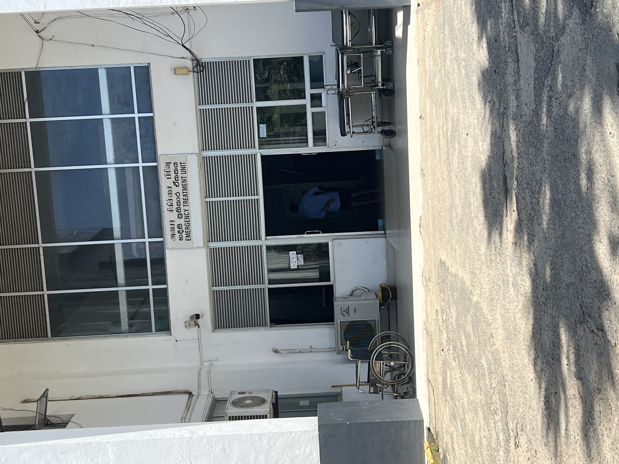 Here is a detailed description of the image:
**Location:** The image shows the exterior of a building, likely a hospital or medical facility in Unknown, Unknown, with a sign indicating an "Emergency Treatment Unit."
**Environment:** The building is constructed from a light-colored material, possibly concrete or plaster. The immediate surroundings are paved with concrete. Sunlight casts shadows on the right side of the image, suggesting a sunny day.
**Objects:**
* **Building Facade:** Features a combination of glass windows and grated panels.
* **Sign:** Has text in English and another language, identifying the area as an "Emergency Treatment Unit."
* **Medical Equipment:** Two wheeled stretchers are parked outside the entrance.
* **Air Conditioning Units:** Two air conditioning units are mounted on the exterior wall.
* **Wheelchair:** A wheelchair is present near one of the AC units.
* **Electrical Wires:** A bundle of electrical wires run along the top of the building.
**People:**
* A person wearing a blue shirt is visible inside the emergency unit.
**Actions/Interactions:**
* The person in the blue shirt is indoors, presumably receiving or administering medical care.