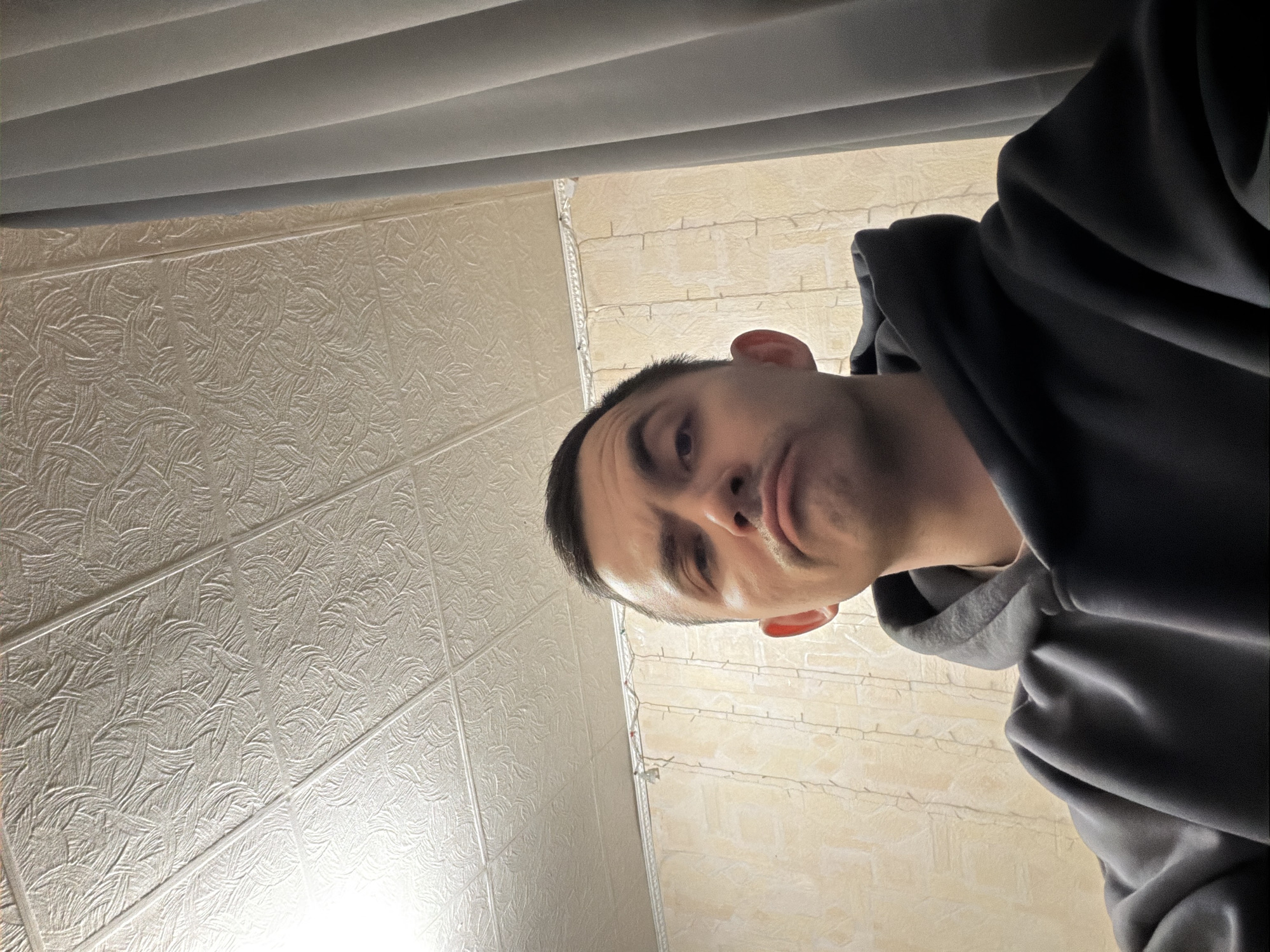 The image is an extreme close-up, low-angle selfie of a person with short dark hair, fair skin, and dark eyes. They are wearing a dark gray hooded sweatshirt. The background consists of a textured white ceiling with repeating pattern tiles and a horizontal gray blind at the top of the frame. The individual's face is angled towards the camera, displaying a slightly puckered expression. There is no indication this image was taken in Izhevsk, Russia.