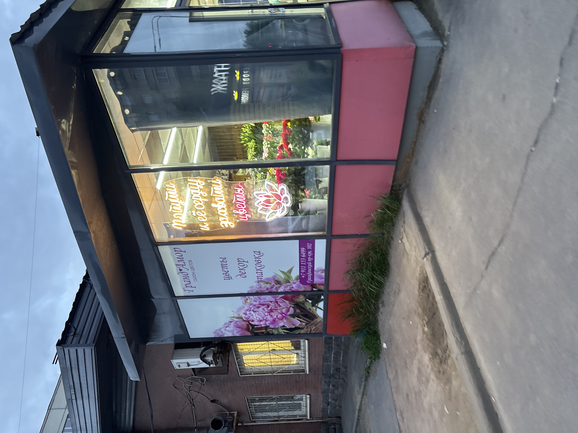 The image shows the storefront of a flower shop named "Гранд Амор". The sign above the shop window advertises flowers, decor, and packaging. There is a phone number listed, +7 911 153 6499, and an address: Гражданский пр-кт, 102. The shop's name, "Гранд Амор," and the types of products it offers ("цветы декор упаковка") are written in Russian. Inside the shop window, there are neon signs that display "Мотити и её сердцу захватий цветы" and a lotus flower design. There are flowers visible inside. The shop is situated on a street with other buildings visible in the background.