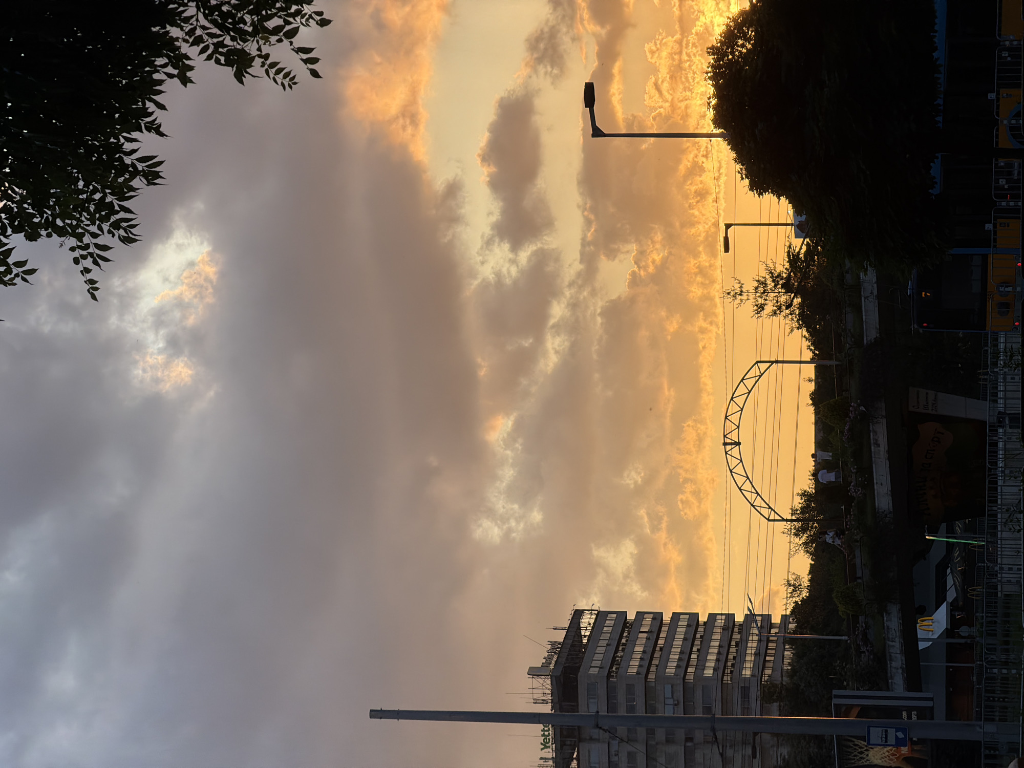 The image depicts an urban scene at sunset in Sofia, Bulgaria, specifically around the National Palace of Culture (NDK) at the intersection of Bulgaria Blvd and Vitosha Blvd. The sky is largely covered by clouds, displaying a warm palette of grey, pink, orange, and golden yellow, with the brightest light concentrated towards the horizon on the right. Dark green foliage from an unseen tree frames the upper right corner.
On the left, a tall, multi-story building, identifiable as the structure at Bulgaria Blvd. 1, is visible. The top section of this building features signage including "Yettel" in green text and "Media Space" in white text, and appears to have scaffolding or structural elements on its roof. A slender, grey pole stands vertically in the foreground, adjacent to the building.
The mid-ground reveals an elevated pedestrian walkway or bridge, upon which several human figures are discernible, standing or moving. Above this structure, a prominent, dark metal arch framework is present, characteristic of the NDK overpass. Below the elevated structure, a faint McDonald's logo is visible, along with partially visible Cyrillic graffiti on a concrete wall, which reads 'ВЛИЯНИЕ СПОРТ' (Influence Sport). Numerous overhead electrical wires extend horizontally across the mid-ground.
The right side of the image is dominated by the dark silhouettes of dense trees and foliage. Two street lights with dark, bent fixtures are visible against the bright sky, one prominently positioned on the far right and another smaller one further to the left. In the lower right, a yellow tram or bus is partially obscured by dark metal fencing and vegetation. The immediate foreground is delineated by a dark metal fence or barrier system.