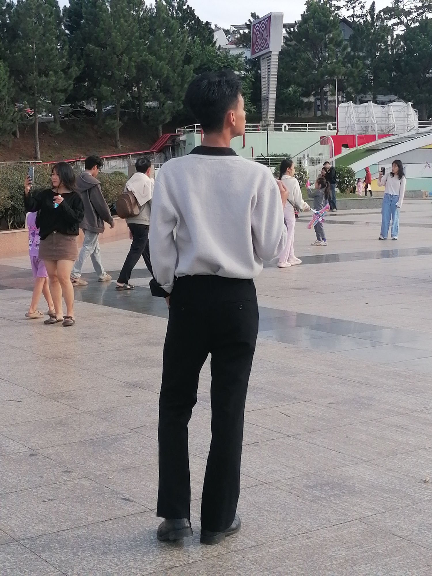 The image depicts a public outdoor area in Da Lat, Vietnam, likely a plaza or promenade with a paved ground surface. In the foreground, a young man is viewed from behind, wearing a light gray sweater and black trousers. To his left, a young woman in a black long-sleeved shirt and a brown skirt holds up a smartphone, appearing to take a picture or video. Behind her, a young girl in a purple t-shirt and shorts walks away from the camera. Further in the background, several other individuals are present, including a man in a gray hoodie and backpack, a woman in a white top and light pink pants, and a woman in a white long-sleeved shirt and blue jeans. There are trees lining a sloped embankment in the background, and a large, colorful sign with the word "GO!" is visible in the upper part of the frame. Structures resembling buildings and amusement park features are also present in the distance. The lighting suggests it is daytime, possibly late afternoon, with soft shadows cast on the ground.