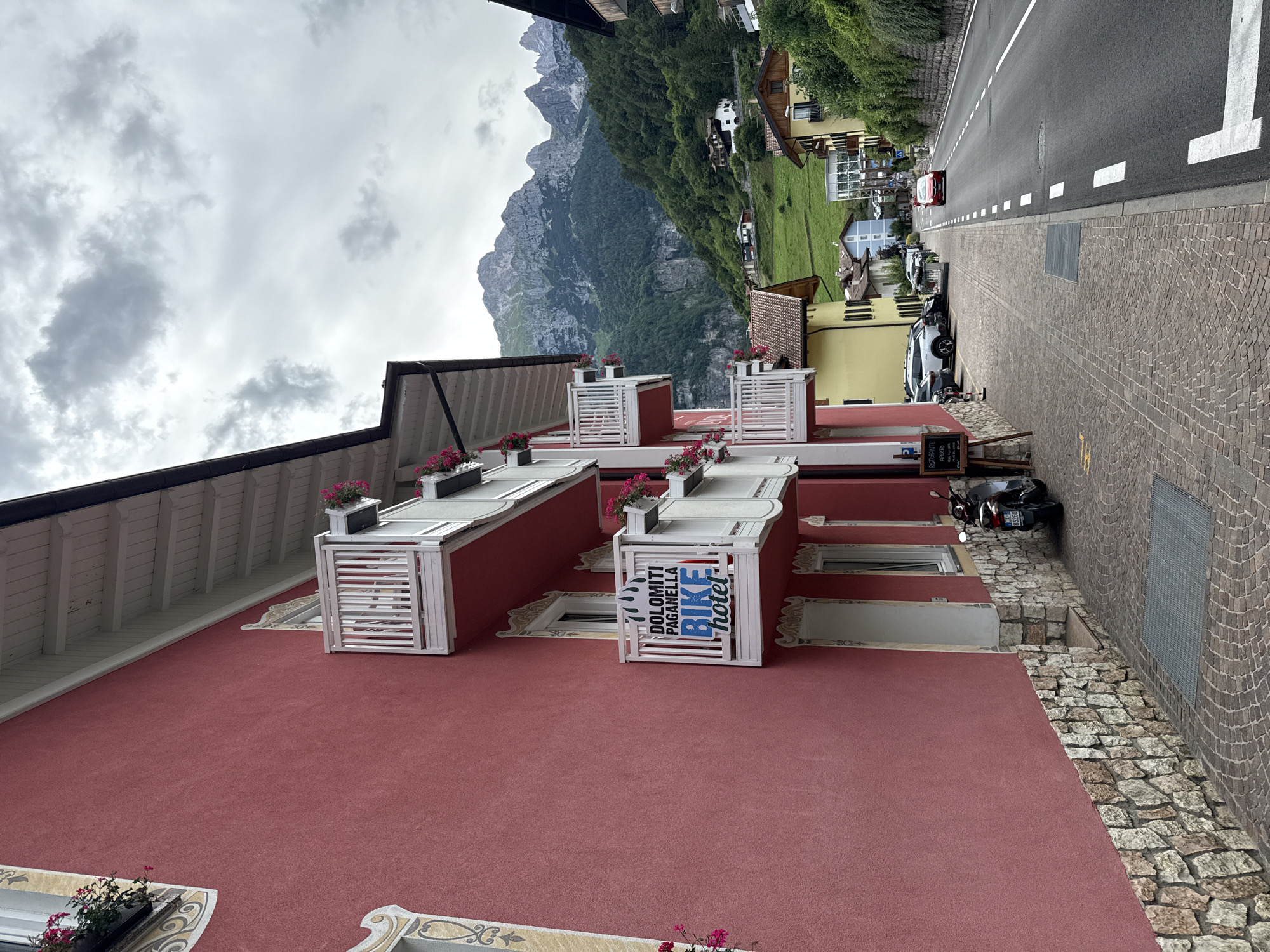 This image captures a street scene in Molveno, Italy, characterized by its mountainous backdrop and traditional architecture. The foreground is dominated by the exterior of a red building, possibly a hotel, with white decorative elements and window frames. Several planters with pink flowers adorn the facade, adding a touch of color. A prominent sign is visible on the building, displaying "DOLOMITI PAGANELLA BIKE hotel" in blue and white lettering.
To the right of the building, a cobbled street runs alongside it, with a road further to the right, marked with white lines. Several vehicles are parked along the street, including cars and a motorcycle. In the background, the lush green slopes of a mountain rise dramatically, partially obscured by a cloudy sky. The presence of houses and trees interspersed on the mountainside suggests a village nestled within the natural landscape.
The overall impression is that of a serene, perhaps slightly overcast day, conducive to outdoor activities like cycling, as suggested by the "BIKE hotel" sign. The architecture and the natural scenery point to a picturesque Italian alpine setting. There is no explicit activity taking place other than general street life and the implied operation of the hotel.