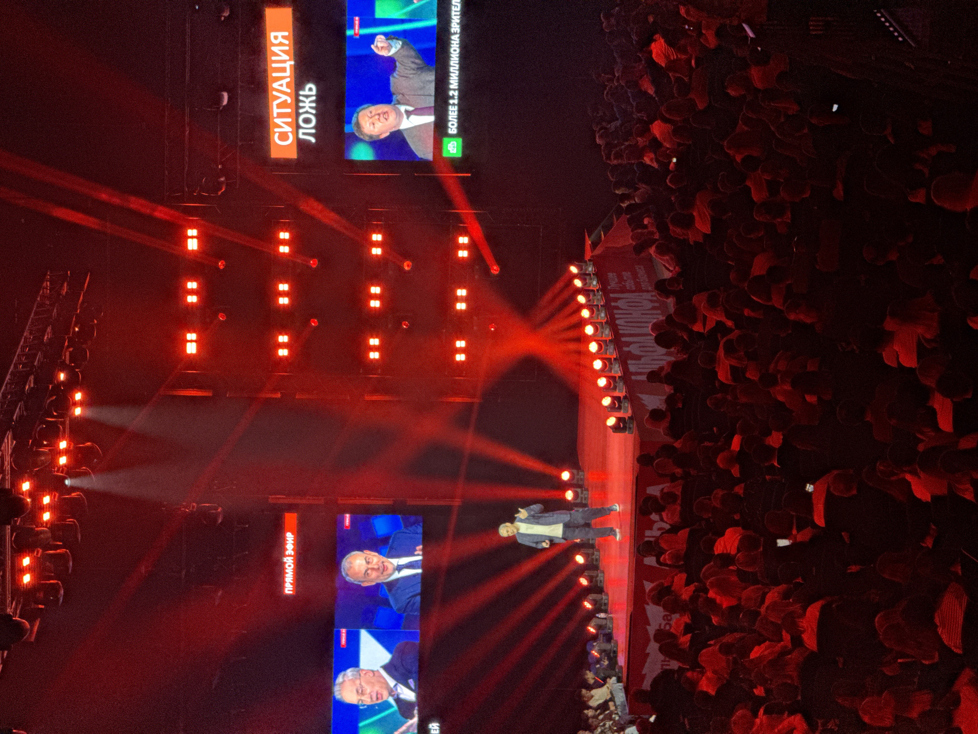 The image depicts a lively event at a stadium or large venue, bathed in dramatic red lighting. In the foreground, a stage is set up with multiple screens displaying close-ups of men in suits. One screen shows a man gesturing with his fist raised, while another displays a man speaking. Red spotlights crisscross the stage, illuminating the smoke or haze in the air.
A man in a grey jacket and dark pants stands on the stage, facing the audience and gesturing with his hands. He appears to be speaking or presenting. To the right of the stage, a massive crowd of people is visible, packed together and illuminated by the red lights. Many in the crowd are holding up their phones, suggesting they are recording or photographing the event. The atmosphere appears energetic and engaging, with a sense of anticipation or excitement.
On the stage, several large banners with text are visible. One banner on the left side of the stage has orange text that reads "СИТУАЦИЯ" (SITUATION) and below it "ЛОЖЬ" (LIE). Another banner on the right has white text that appears to be in Russian. On the screens, some text is also visible, including "БОЛЕЕ 1.2 МИЛЛИОНА ЗРИТЕ" (OVER 1.2 MILLION SPECTATORS) and "ПРЯМОЙ ЭФИР" (LIVE BROADCAST). The overall scene suggests a political rally, a concert, or a large public address. The time of day is likely evening or night, given the artificial lighting and indoor/covered setting.