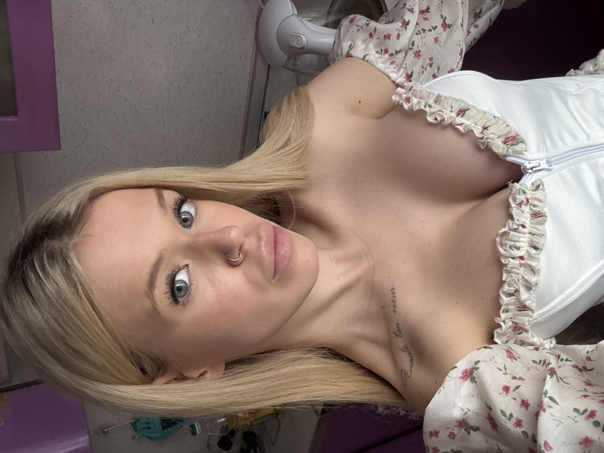 A young woman with blonde hair and blue eyes is shown in a close-up, selfie-style shot. She is wearing a white off-the-shoulder top with a floral print and a ruffled neckline. A zipper is visible at the front of the top. She has a nose ring and a tattoo on her left collarbone. The background appears to be indoors, with a gray, textured wall and some purple elements visible. There is also a spray bottle with a gray nozzle on a shelf in the background. No specific location in Saint Petersburg, Russia can be identified from the image.