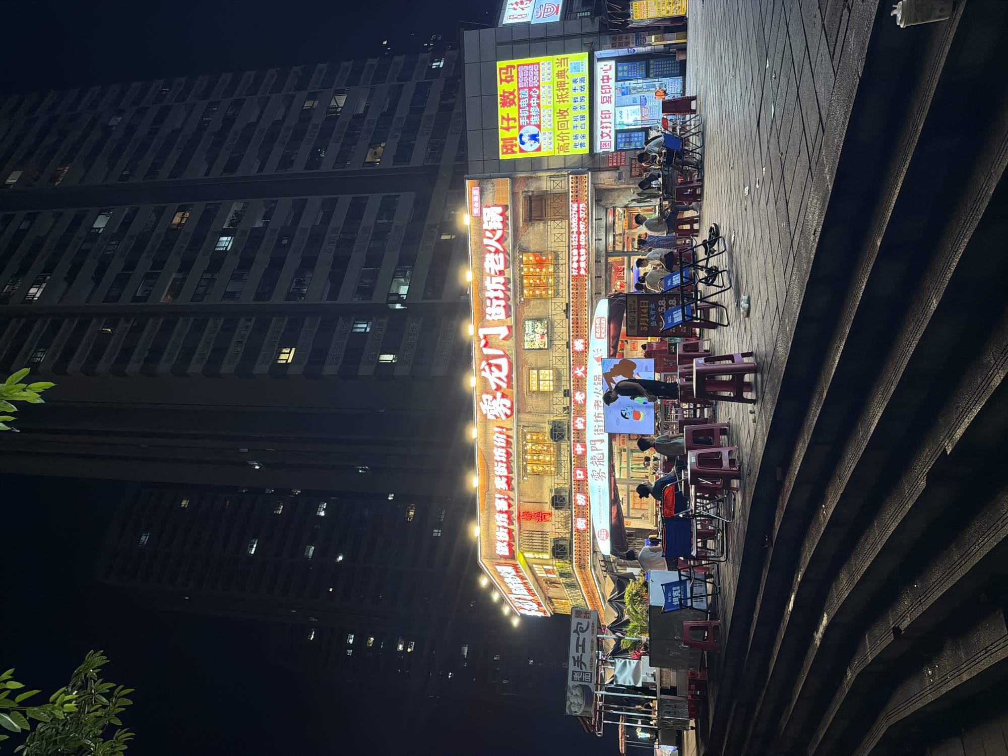 The image shows a night scene in Chongqing High-tech Zone, China, featuring a brightly lit commercial street lined with restaurants and shops. A tall apartment building looms in the background. People are sitting at tables outside a restaurant called 雾龙门 (Wu Long Men), which specializes in hot pot. Neon signs in Chinese characters are prominently displayed above the storefronts. The street is paved with dark grey stone tiles and has a set of stairs along one side. There are various objects, including chairs, tables, and advertising signs, scattered throughout the scene. Some people are eating and talking, while others are walking by. The overall atmosphere is lively and bustling.