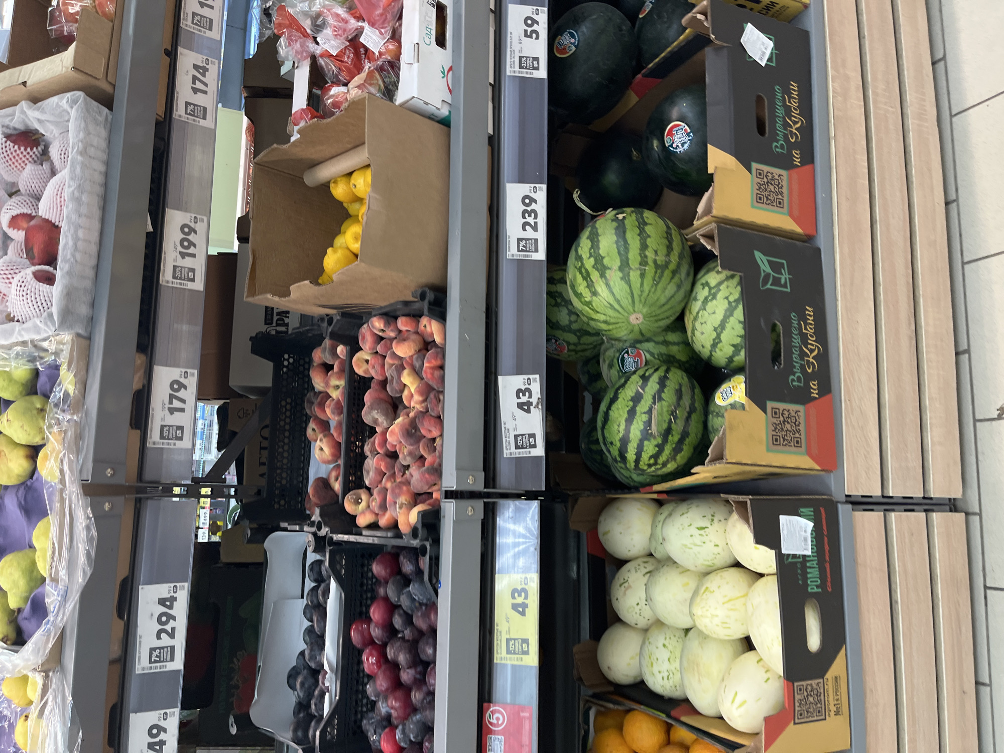 The image shows a produce aisle in a supermarket, likely in Moscow, Russia, based on the Cyrillic text visible on product labels and signs. The shelves are stocked with various fruits and vegetables.
In the top shelf, there are several types of fruit, including pears, plums, and peaches. Some are displayed in plastic containers, while others are in cardboard boxes. Signs with prices in rubles are visible above these items.
The middle shelf features more produce, including more peaches, lemons in a cardboard box, and what appear to be large red fruits in plastic packaging.
The bottom shelf displays a variety of watermelons and light-colored melons in cardboard boxes. Some of the watermelon boxes have QR codes and Cyrillic text indicating they were grown in a specific region.
The overall environment is a well-lit supermarket aisle with commercial shelving units. No people are visible in the image. The focus is on the arrangement and display of the produce for sale.