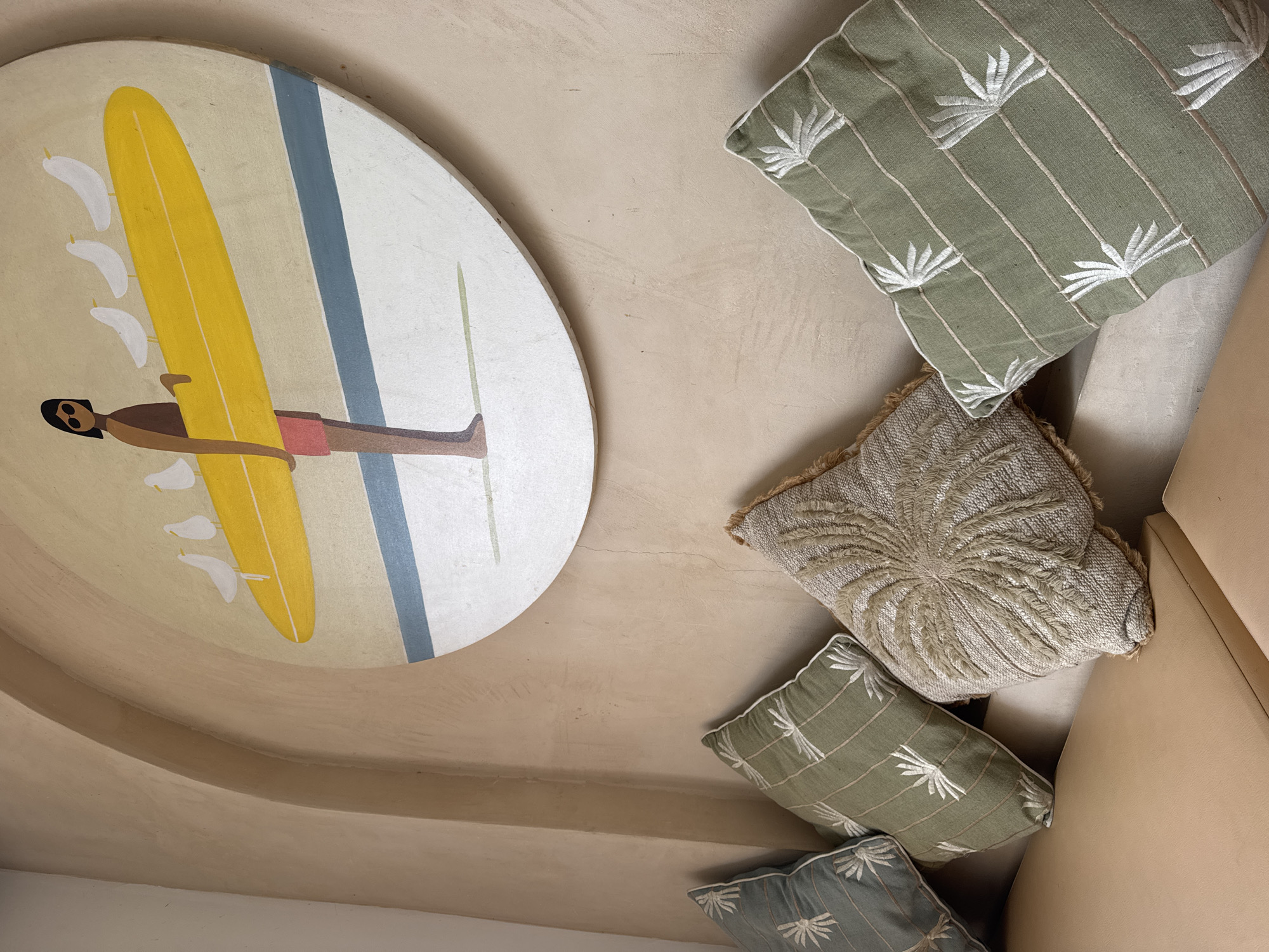 The image depicts an indoor scene featuring a large, circular painting mounted on a textured, light-brown wall. The painting is in a minimalist style, showing a person in a brown swimsuit and red shorts holding a yellow surfboard with several white birds perched on it. Below the surfboard is a blue stripe and a white area, representing water and sand.
In front of the wall, there is a tan-colored sofa or bench. On the sofa, there are several decorative cushions. From left to right, the cushions include: one dark green cushion with a white embroidered palm leaf pattern, a light green cushion with a similar white embroidered palm leaf pattern, a textured, beige cushion with a circular woven design and fringed edges, and another light green cushion with the white embroidered palm leaf pattern.
The wall has an arched alcove or recess behind the painting. The overall impression is of a decorative interior space, possibly a lounge or seating area. The scene is well-lit, and the focus is on the artwork and the cushions. The environment appears clean and well-maintained. The specific location in Kuta Selatan, Indonesia is not visually evident from the image itself.