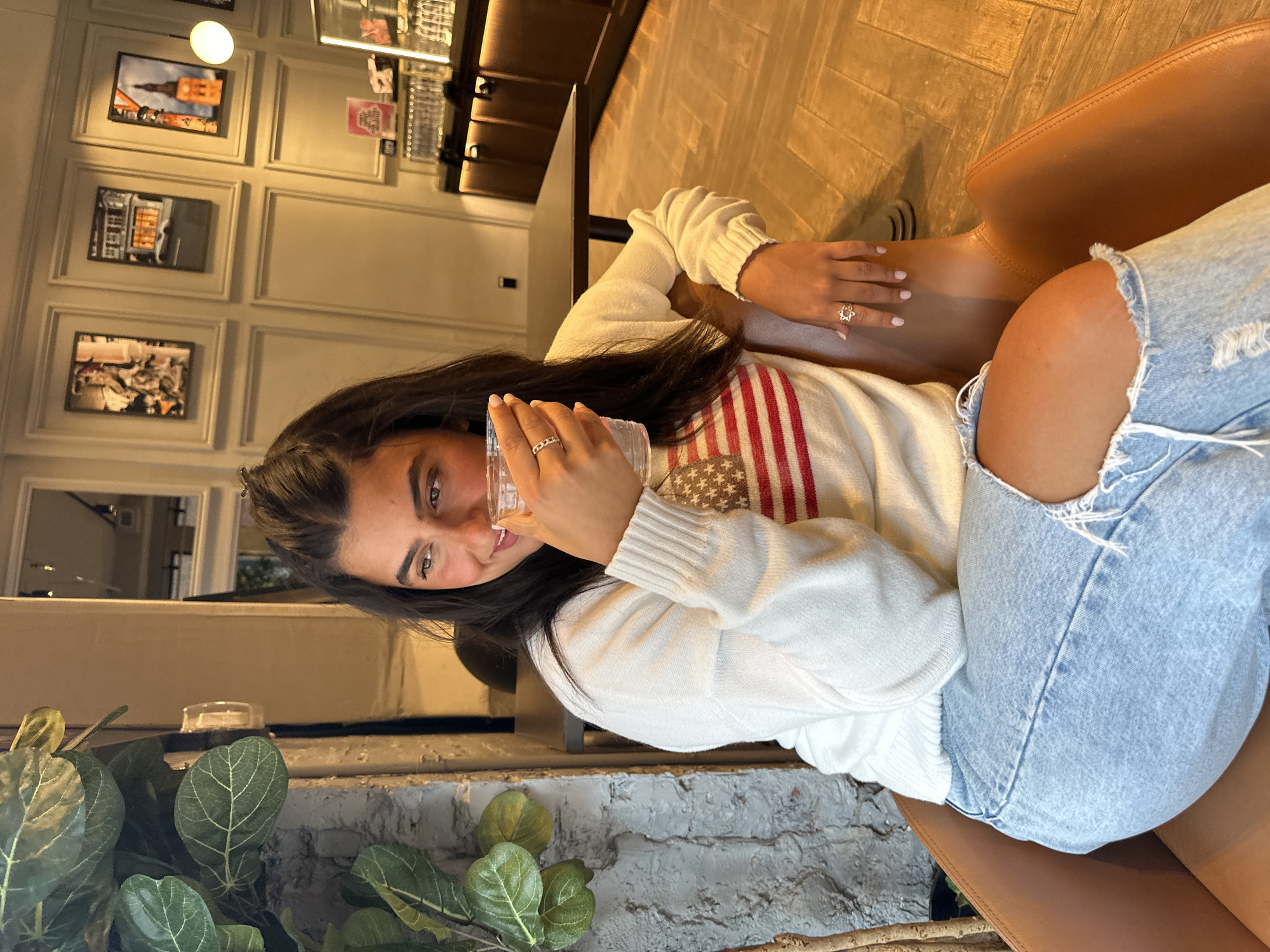 A young woman with long dark hair is captured in a vertical, slightly tilted indoor shot, appearing to be in a cafe, restaurant, or lounge. She is seated on a brown leather chair, looking directly at the camera with a slight, friendly smile. She wears a cream or white long-sleeved knitted sweater featuring a prominent red and blue striped or star-patterned design on the front, possibly resembling a flag. She also has on light blue ripped jeans. Her fingernails are painted a light color, and she wears two rings: a band on her left ring finger and a more decorative, possibly floral or star-shaped, ring on her right ring finger. She holds a clear glass or cup in her right hand, near her mouth.
The background features a light-colored wall adorned with multiple framed pictures depicting urban scenes or buildings. A dark table is visible to her right. In the foreground, a large green plant with prominent leaves is positioned to the left, set against a brick-textured wall. The flooring is wooden. The lighting suggests an indoor setting, likely evening or late afternoon, with artificial light sources. The overall atmosphere is relaxed and casual. No discernible text is visible on any objects or in the background. The location is understood to be in New York, United States of America.