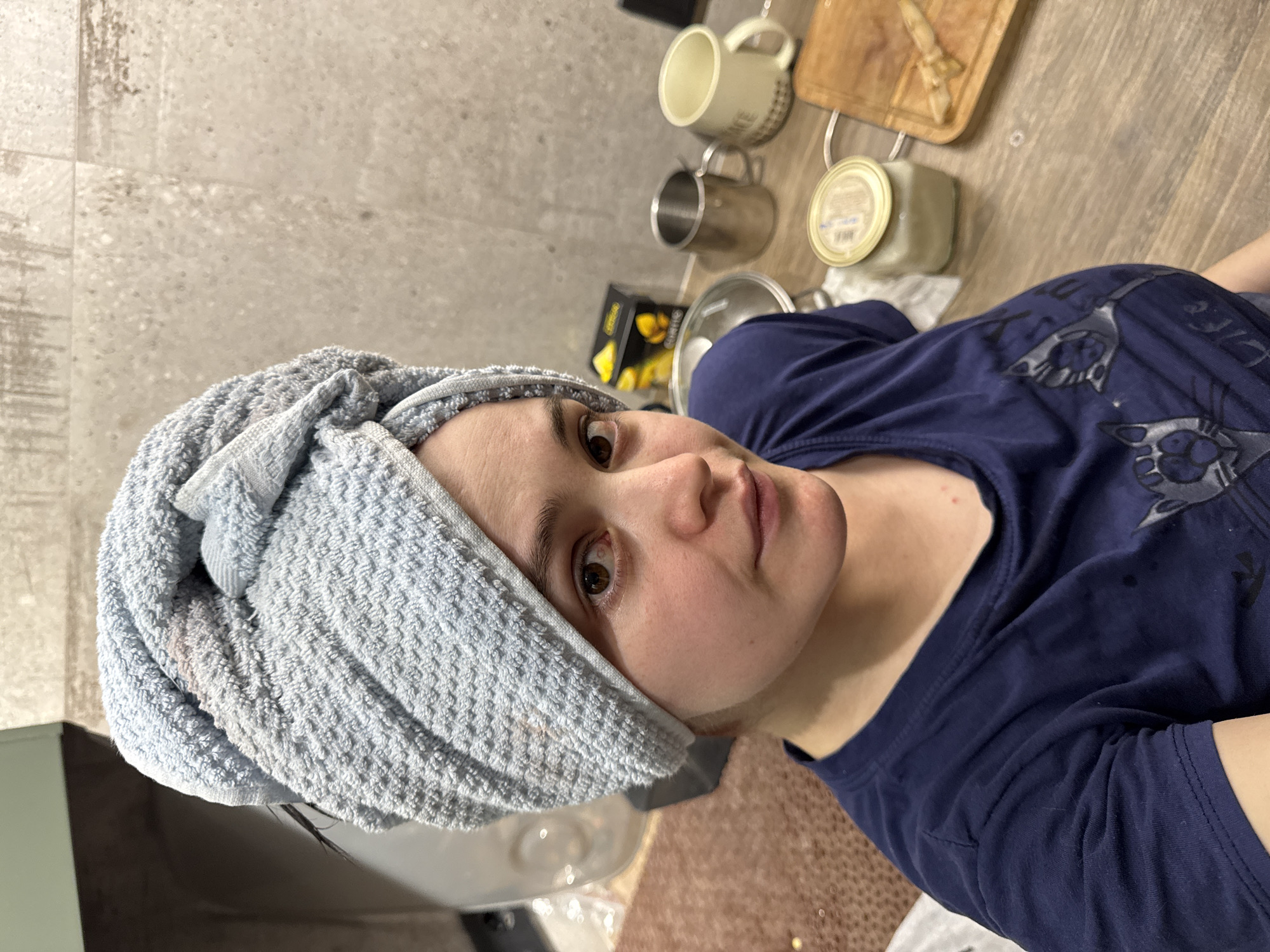 An adult individual, likely female, is depicted in a portrait-style image rotated 90 degrees counter-clockwise. She looks directly at the camera with a neutral expression, her eyes appearing wide and slightly reddish, possibly from recent washing. Her hair is wrapped in a light blue, textured towel, which shows a distinct reddish-pinkish stain on the side near her temple. A few dark strands of hair are visible peeking out from under the towel. She is wearing a dark blue t-shirt with a faded, abstract grey or silver pattern. Minor red spots are noticeable on her neck and upper chest.
The setting appears to be a domestic kitchen or utility room. The background features a light-colored, textured tiled wall. Below the wall, a countertop or floor with a wood-grain pattern holds several items: a light ceramic mug, a metallic mug, a wooden cutting board with what resembles garlic cloves, a glass jar with a metallic lid and a label (indistinct), and a black box with yellow details, possibly bearing the word "Lemon." The lighting suggests indoor artificial illumination. The image captures a candid moment, perhaps after a shower or hair wash. The location context for this scene is Izhevsk, Russia.