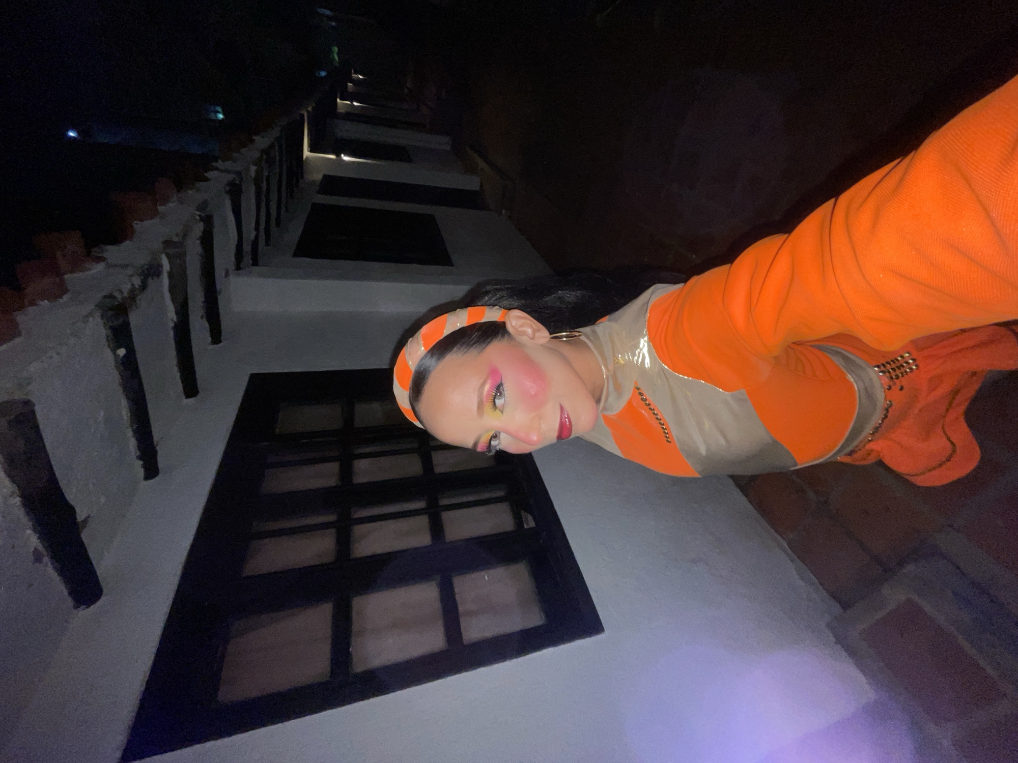 A person is standing in a dimly lit outdoor walkway at night. The person is wearing an orange and beige outfit with a matching headband and has bold makeup. The walkway appears to be lined with white buildings that have dark-framed windows and exposed wooden beams under the roof overhangs. The ground is paved with brick. The lighting suggests the scene is in Porlamar, Venezuela, likely a hotel or guesthouse with a rustic architectural style.