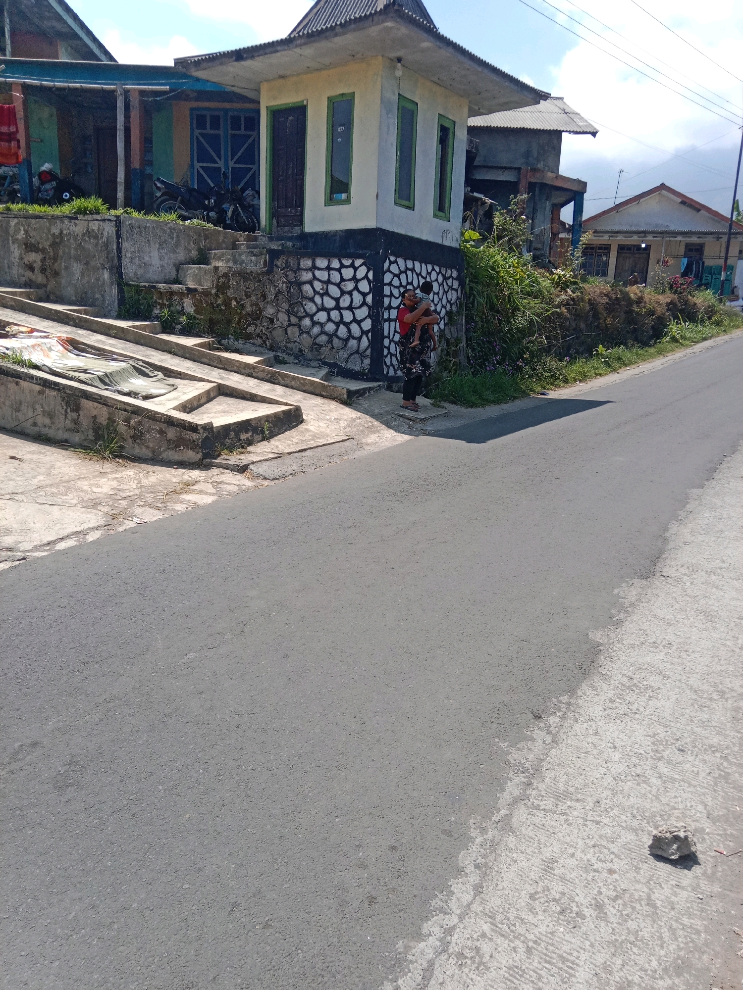 A wide-angle shot captures a sunlit, rural Indonesian street in Adipuro. The asphalt road dominates the foreground and midground, with a concrete embankment running alongside it on the left. On the embankment, there are several concrete steps and a covered structure, possibly for storage.
In the background, on the right side of the road, a small, tan-colored building with a dark-tiled roof stands prominently. It features two tall, narrow windows with green frames and a black-and-white patterned wall at its base. To the left of this building, more structures and vegetation are visible.
A woman in a red shirt and patterned skirt is standing on the concrete area in front of the small building, holding a child. Behind them, the scene continues with more houses, a motorcycle parked on a grass-covered area, and lush green vegetation. The sky above is clear with scattered clouds. The overall impression is a glimpse into daily life in a Southeast Asian village.