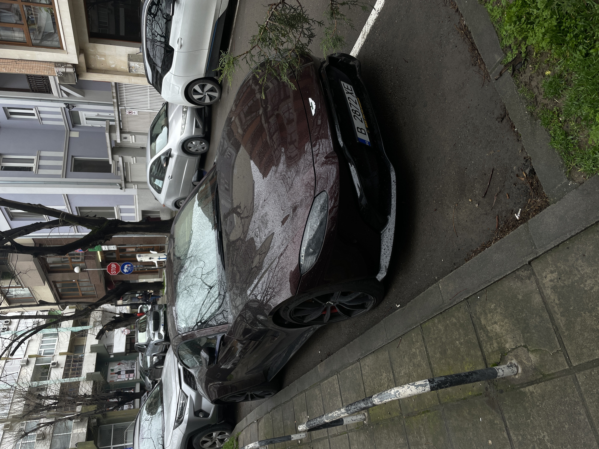 The image shows a street scene in what appears to be an urban area. A burgundy car with a license plate "B 2872 XE" is parked along the curb. Other cars, including a white BMW i3, are parked nearby. Buildings, possibly apartments, are visible in the background, with trees interspersed along the street. A "STOP" sign is present. The setting is likely Varna, Bulgaria, based on the license plate country code. The scene appears to be slightly wet, possibly after rain. There are no people prominently visible. The focus is on the parked cars and the urban environment.