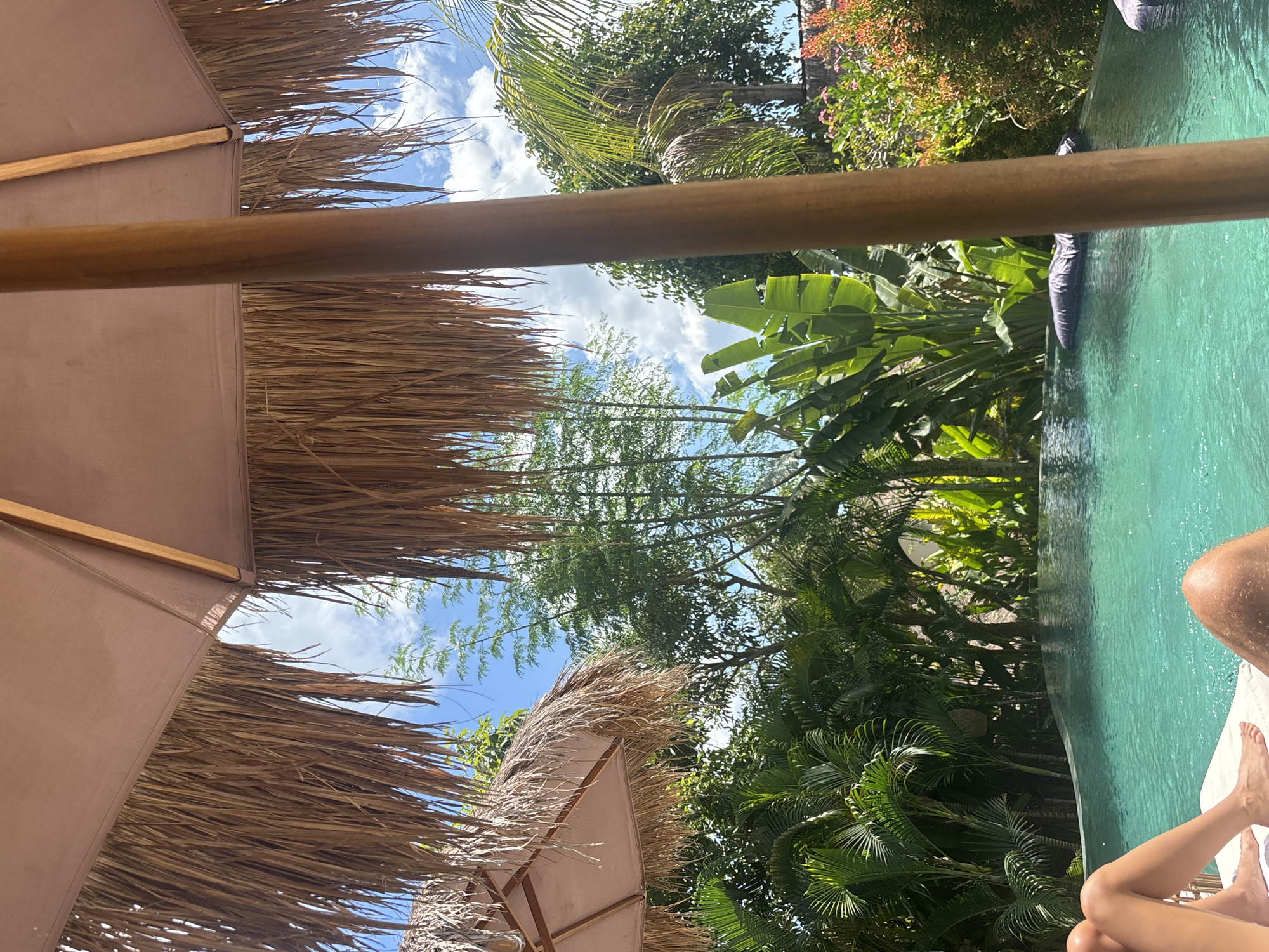 The image depicts a scene in Kuta Selatan, Indonesia, featuring a poolside view under a straw-thatched umbrella. Two individuals are partly visible in the lower right corner, showing legs and feet resting on a white surface, likely a lounge chair. A swimming pool with turquoise water occupies the right side of the frame. Lush tropical vegetation, including palm trees and banana leaves, surrounds the pool area. Two more straw-thatched umbrellas are partially visible in the bottom left of the image. The sky with scattered clouds is visible through the vegetation.