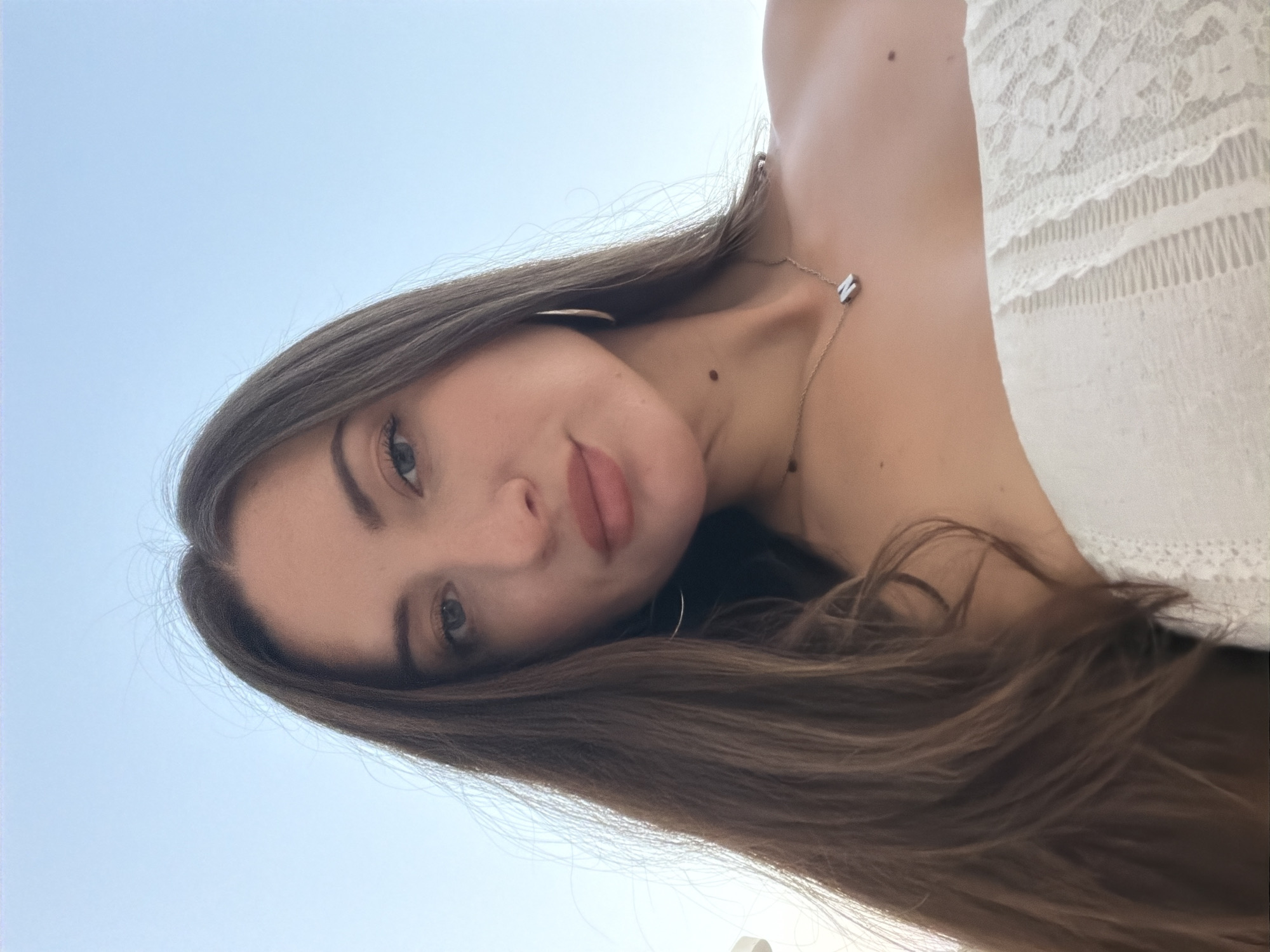 This is a head and shoulders selfie of a woman with long, dark brown hair and fair skin. She is wearing a white, strapless, lace top and a delicate necklace with a rectangular pendant. The background is a clear blue sky, suggesting an outdoor setting in Sharm El Sheikh, Egypt, likely during daylight. The woman is looking directly at the camera with a neutral expression. The lighting is soft and natural.