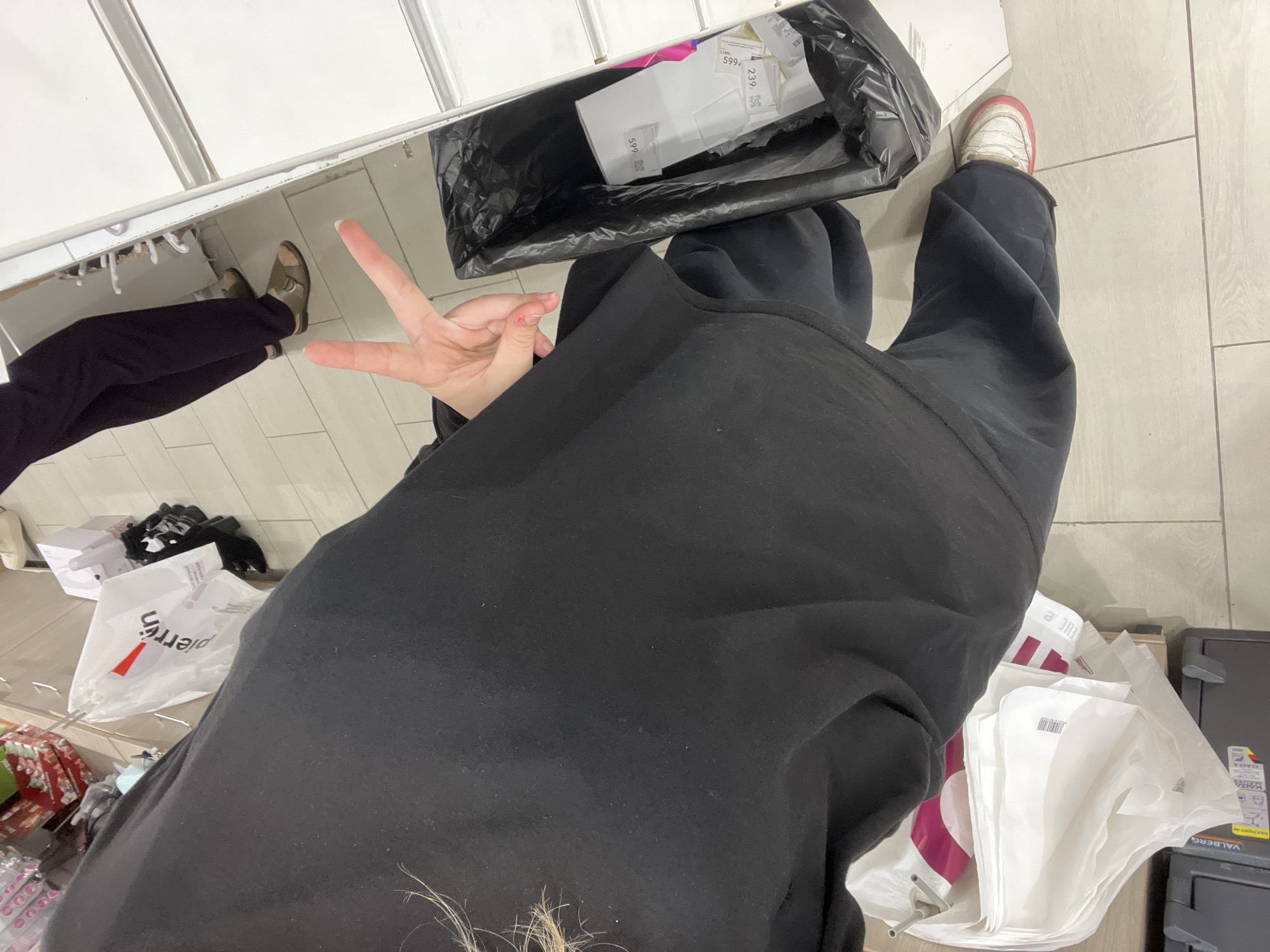 A person, viewed from a high angle, is standing in front of a metal counter and a tiled floor. The person is wearing a black sweatshirt and black pants, with white and pink sneakers on their left foot. Their right hand is raised, fingers extended in a peace sign. To the left of the person are several white plastic shopping bags. In front of the person is a black trash bin with white paper items inside. To the right, a reflection in a mirror shows the person's legs and feet, as well as another person's legs wearing dark pants and sandals. The environment appears to be an indoor retail setting, possibly a store. There is no visible indication of the location being Kokkorevo, Russia.