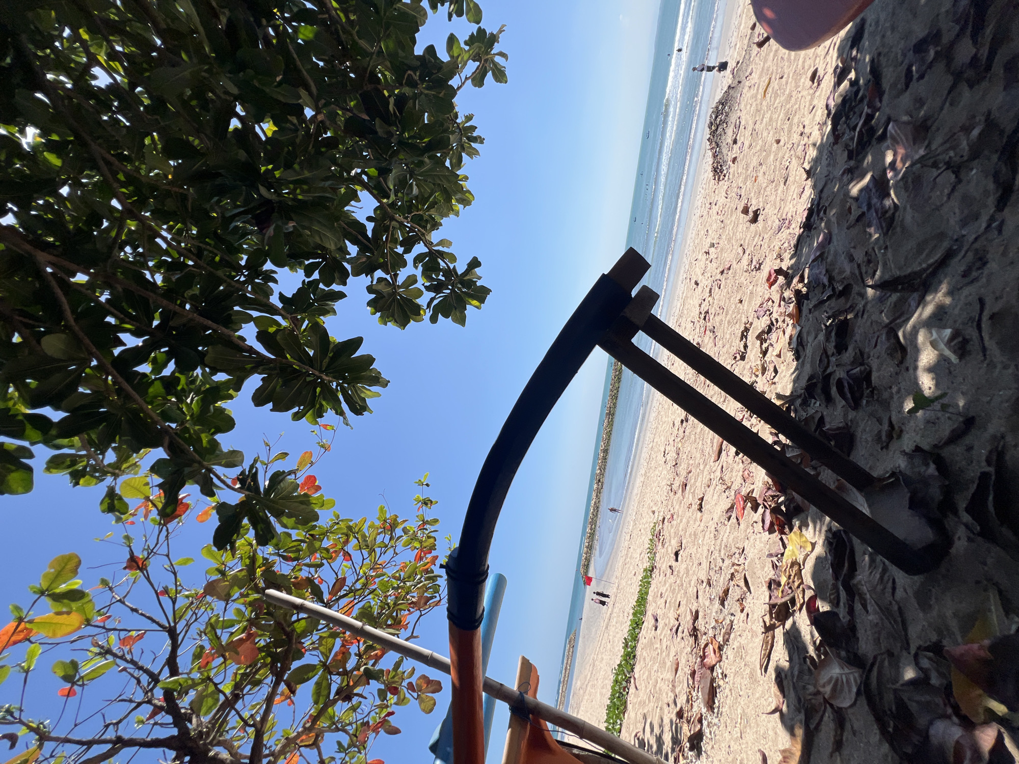 A low-angle shot captures a beach scene in Kuta, Indonesia, from beneath the shade of a leafy tree. The foreground is dominated by fallen leaves and sand, with the dark, curved arm of a beach chair or lounge intruding from the lower left. Beyond this, the sand stretches to the ocean, dotted with a few scattered people and a small red flag. The water is calm, with gentle waves lapping the shore. A stone breakwater extends into the sea in the mid-ground, and a clear blue sky fills the upper portion of the frame. The foliage of the tree, with some leaves showing autumn colors, drapes over the scene, creating a dappled sunlight effect.