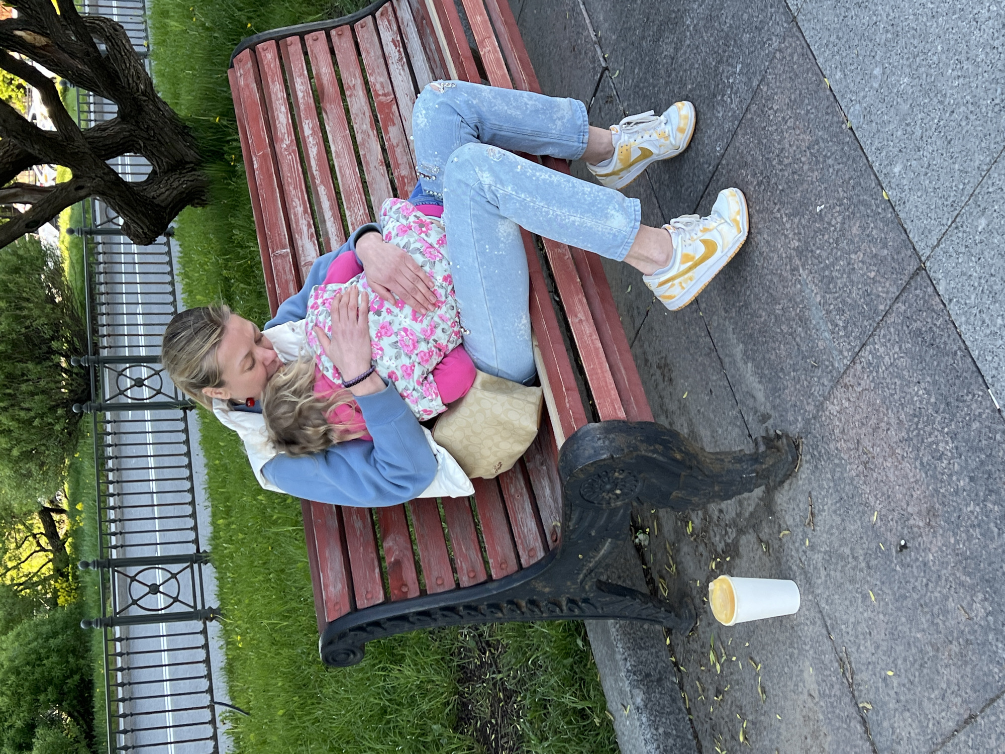 A woman is lying on a red park bench with a young child in her arms. The woman has blonde hair pulled back, wearing a light blue sweatshirt, white vest, and light blue jeans. The child is wearing a pink jacket with a floral pattern. The bench is situated on a gray paved surface next to a grassy area with trees and a black metal fence in the background. A discarded white cup with liquid inside is on the pavement near the bench leg. The location is likely a park or public area in Saint Petersburg, Russia, due to the characteristic appearance of the park benches, fencing, and architecture.