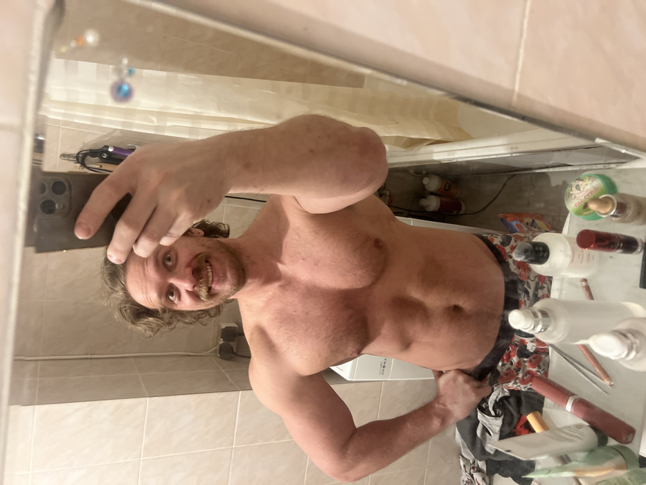 A shirtless man with a mustache is taking a selfie in a bathroom mirror. He's holding his phone in his left hand, showing off his physique. The bathroom is visible in the reflection. It includes various toiletries and a washing machine. The tiles are light-colored.