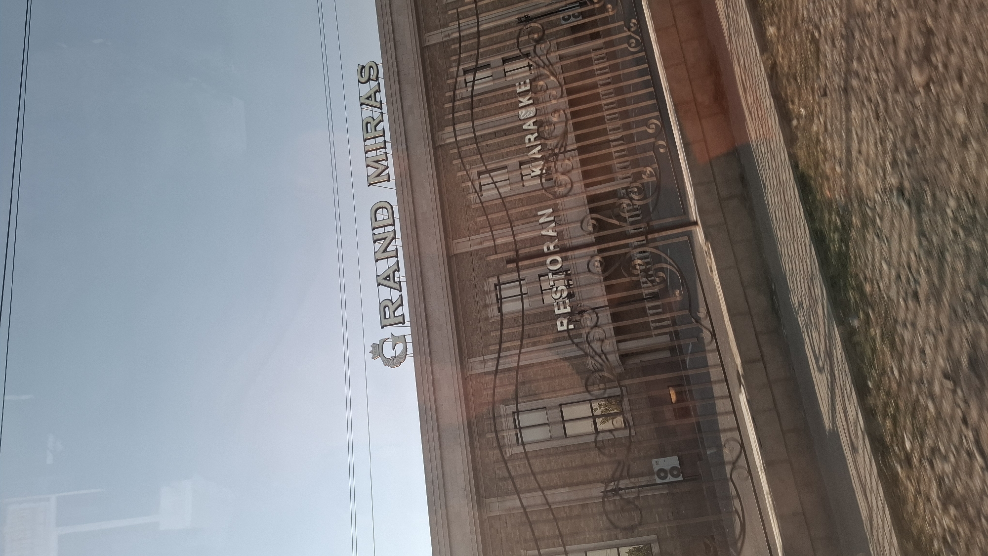 The image shows the exterior of a building with the sign "GRAND MIRAS" prominently displayed on its roof. Below this, on the facade of the building, the words "RESTORAN KARAOKE" are visible. The building appears to be made of brick and has several windows. A decorative metal fence runs across the front of the building. The scene is set in an outdoor environment with a clear sky. Based on the text on the sign and common knowledge of businesses in the region, this is likely the Grand Miras restaurant and karaoke bar located in Shubarssu, Kazakhstan.