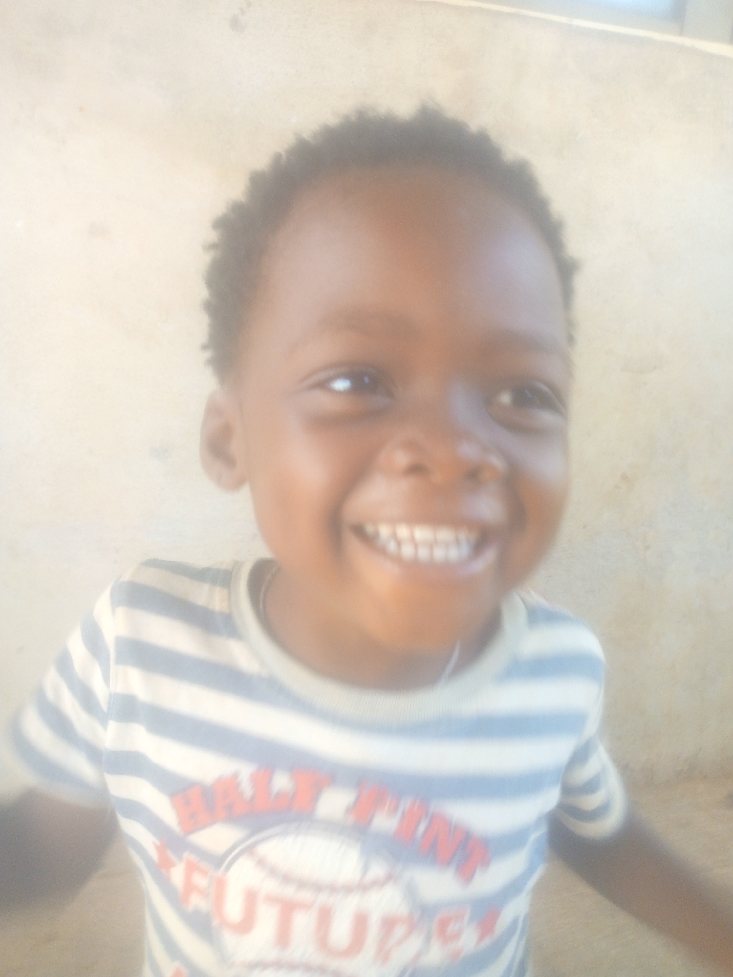 A joyful young child with dark skin and short, curly dark hair is captured in a waist-up, slightly blurry portrait. The child is smiling broadly at the camera, revealing a full set of small teeth and bright, happy eyes. They are wearing a striped t-shirt with alternating white and light blue horizontal stripes. On the chest of the shirt, red text is visible, reading "HALF PINT" at the top and "FUTURE" below it, possibly part of a playful, sports-related graphic like a baseball design. The background consists of a plain, light-colored wall with some faint, irregular marks, suggesting an indoor or sheltered outdoor setting. The scene is brightly and evenly lit, indicative of daytime. The child's expression clearly conveys happiness and cheerfulness.