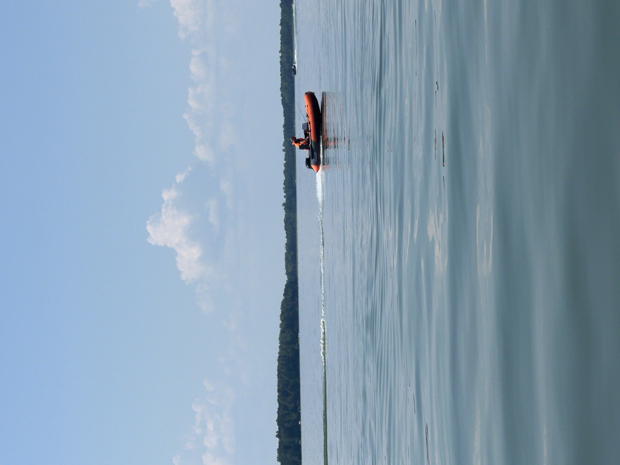 An orange and black inflatable boat with a person wearing an orange life vest is traveling across a calm body of water. The boat is creating a wake. In the background, a line of trees forms the shoreline of what appears to be a lake or large river. The sky is blue with scattered white clouds. The location is identified as Borovoe, Russia.