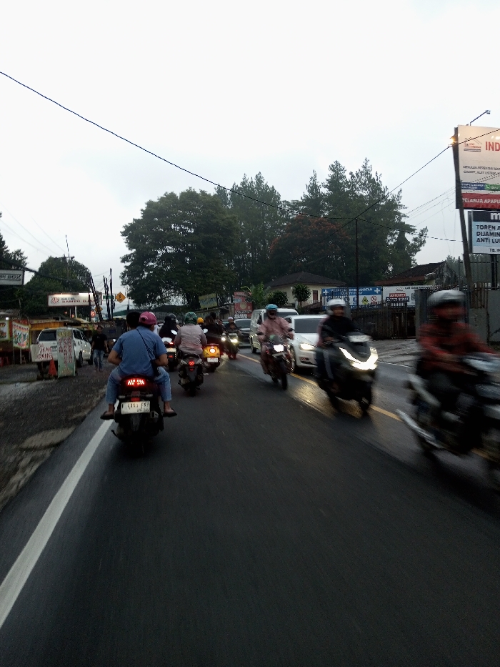 The image depicts a busy street scene in Dawathang_Dorjibi_ Kashingtsawa, Bhutan, under an overcast sky, possibly during or after rainfall. The asphalt road surface appears wet, reflecting ambient light. The scene is dominated by motorized two-wheelers, with multiple individuals traversing the thoroughfare.
In the immediate foreground, a dark-colored scooter is centrally positioned, moving away from the viewer. A person wearing a blue shirt, jeans, and a pink helmet is seated as a passenger, with their feet visible. The rider of this scooter is partially obscured.
Numerous other motorcycles and scooters follow in lanes, extending into the mid-ground. Many riders wear helmets, and some carry passengers. A white passenger car is visible amidst the motorcycles in the mid-ground, heading in the same direction. Several vehicles exhibit motion blur, particularly on the right side of the frame, indicating movement.
Along the left side of the road, a narrow unpaved shoulder or sidewalk is present. Several informal commercial structures and signs are visible, including one white stand displaying "MOTO BOX" and another sign above, partially obscured, with "HOTEL SANTO DA" text. Utility poles with an intricate network of overhead electrical or communication wires crisscross the upper portion of the frame. Lush green trees and dense foliage line the left side, extending into the background.
On the right side of the road, another large utility pole supports multiple billboards displaying various advertisements, some with Indonesian text such as "INDAH" and "BELANJA APAPUN". Beyond the billboards, more dense green trees form a natural backdrop, partially obscuring residential or commercial buildings. The sky is uniformly light grey, indicating an overcast condition and diffuse lighting.