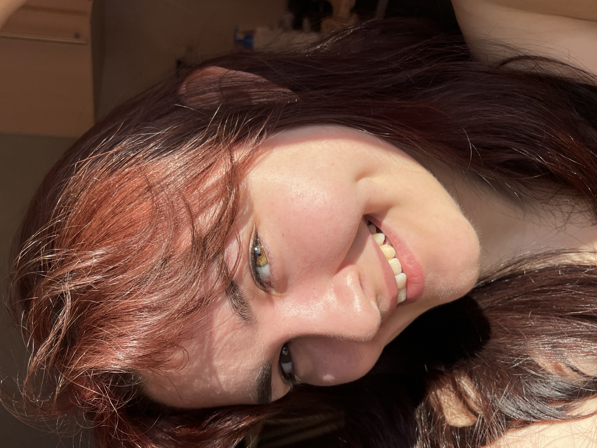 A close-up portrait of a smiling young woman with auburn hair. Her head is tilted to the left. She has fair skin and hazel eyes. Her teeth are visible. The background is out of focus. There is no evidence to suggest the location is in the city of Neftekamsk, Russia.