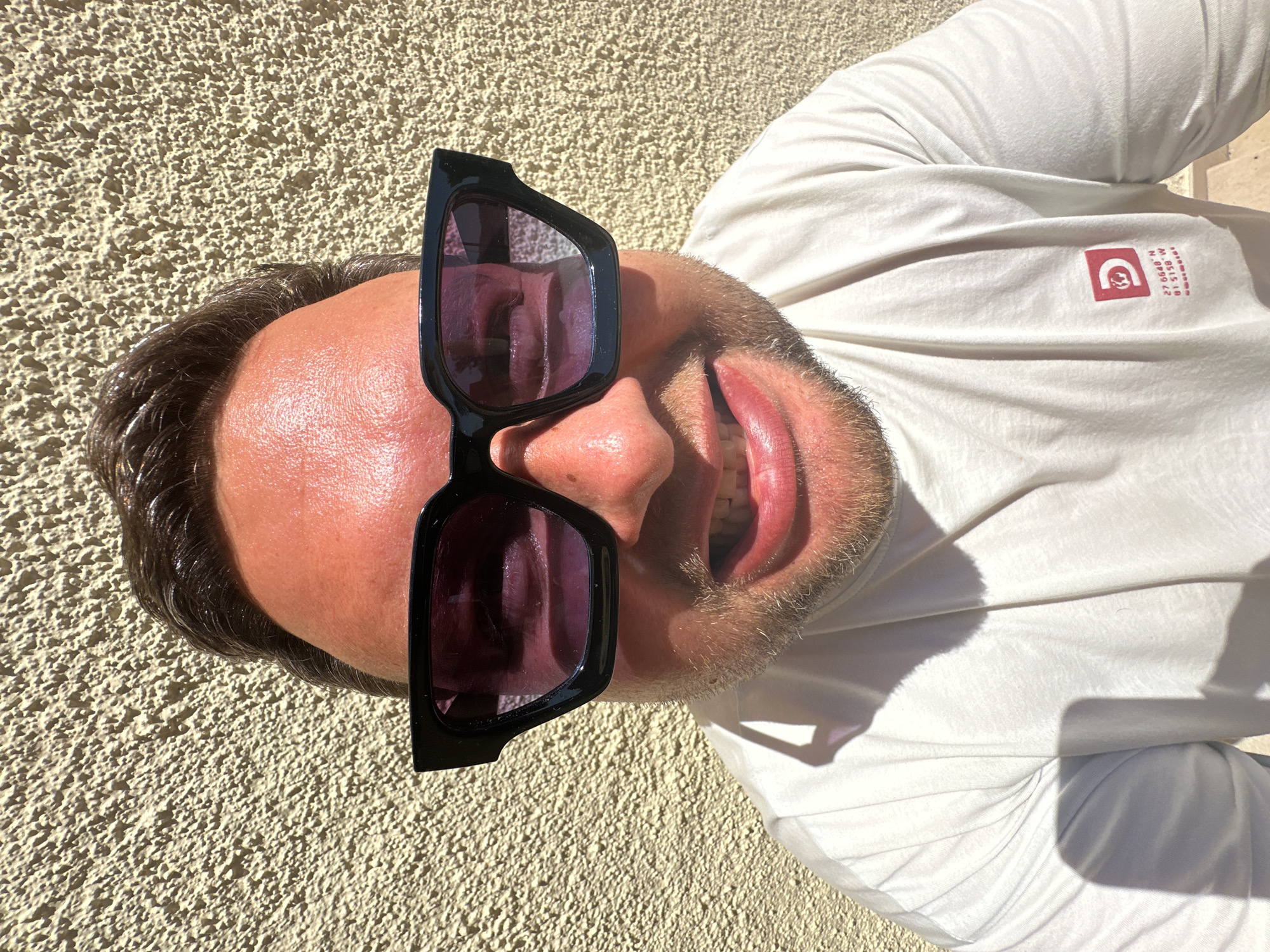 A man with short dark hair and a beard wears sunglasses and a white t-shirt with a logo and text that reads "27.6048° N 81.5158° W" on a sunny day. The background is a textured beige wall, which could be part of a building. The location, Sant Feliu de Guíxols, Spain, is not discernible from the image content.