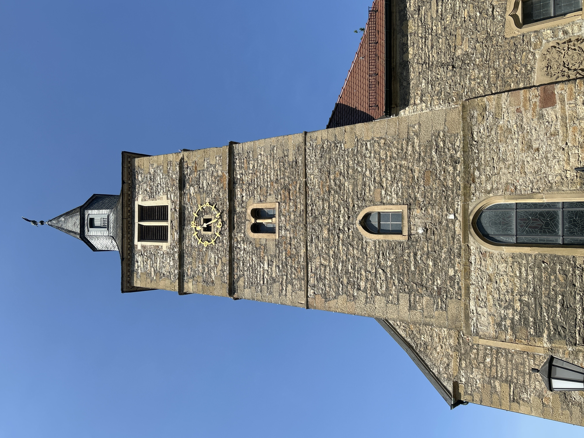 The image shows the stone tower of a church against a clear blue sky in Bad Friedrichshall, Germany. The tower has a peaked roof and features a clock with gold roman numerals. There are several windows of various sizes and shapes on the tower. To the right of the tower, part of the church's red-tiled roof is visible. The stonework of the tower is weathered and shows various shades of brown and gray. A black lantern is visible at the bottom left of the image. There are no people visible in the image. The scene is captured in bright daylight, casting distinct shadows.