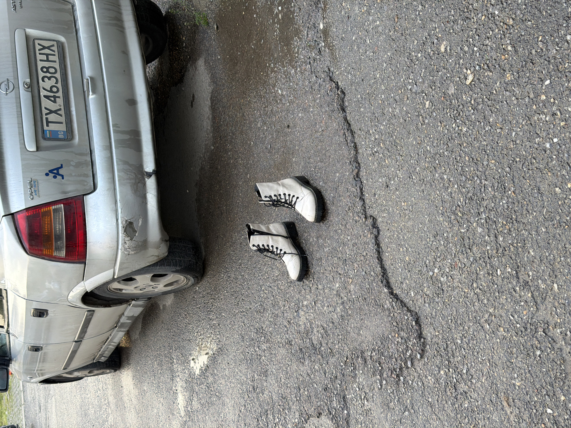 The image shows a silver Opel Astra car, partially visible, parked on an asphalt surface with visible cracks. A pair of white leather boots with black laces are lying on the asphalt next to the car. The car's license plate reads "TX 4638 HX" and has a Bulgarian flag symbol, indicating the location is likely in Bulgaria. There is damage to the rear bumper of the car. The environment suggests an urban setting, potentially in the city of Sofia. No people are visible in the image.