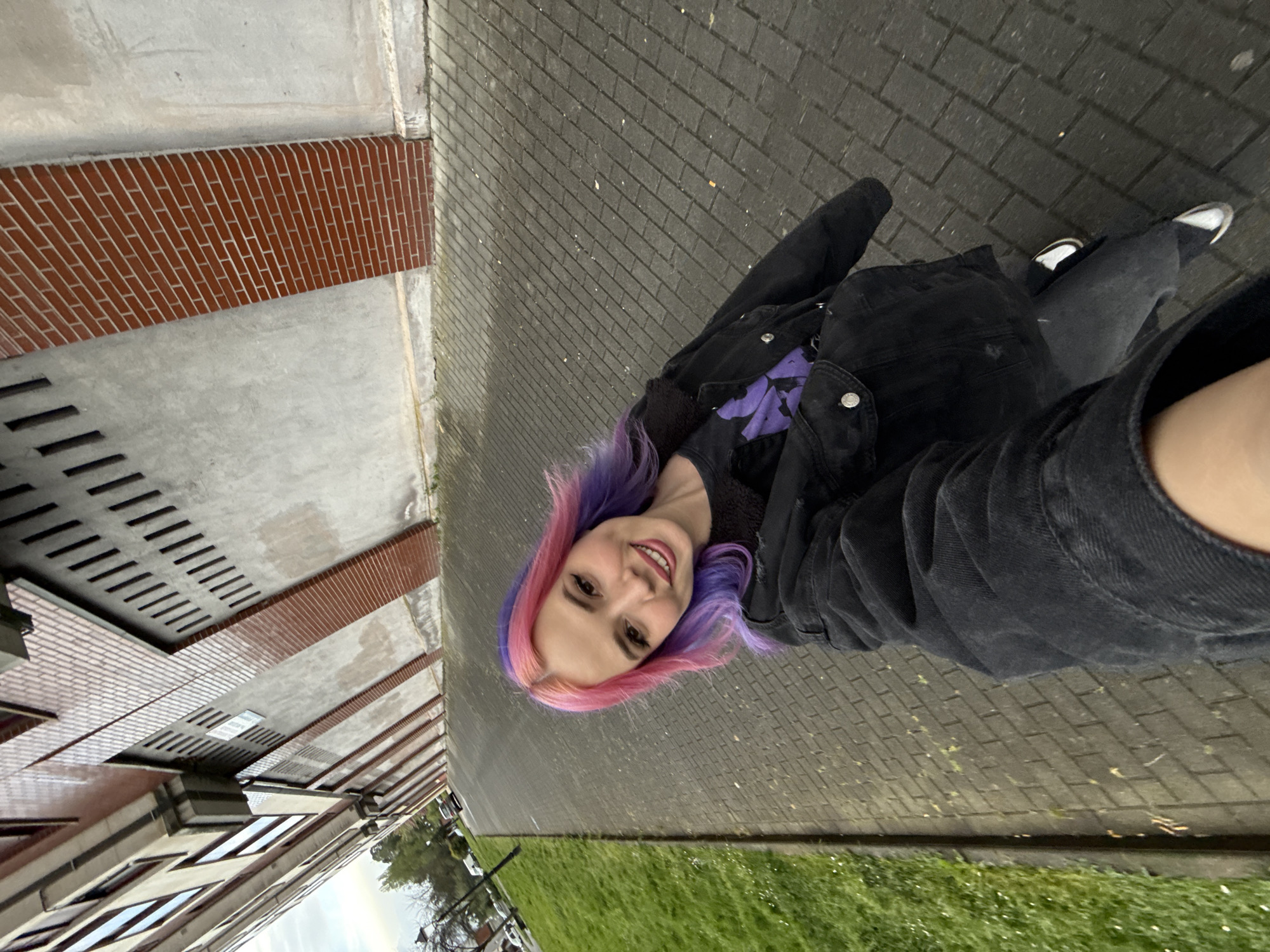 The image shows a young woman with dyed pink and purple hair standing on a paved walkway next to a building in what appears to be El Pontón De Vaqueros, Spain. She is wearing a black denim jacket and a dark-colored shirt with a purple design. The building to her left has a red brick facade with what appears to be window vents. Beside the walkway is an area of grass. The image appears to be taken at an angle.