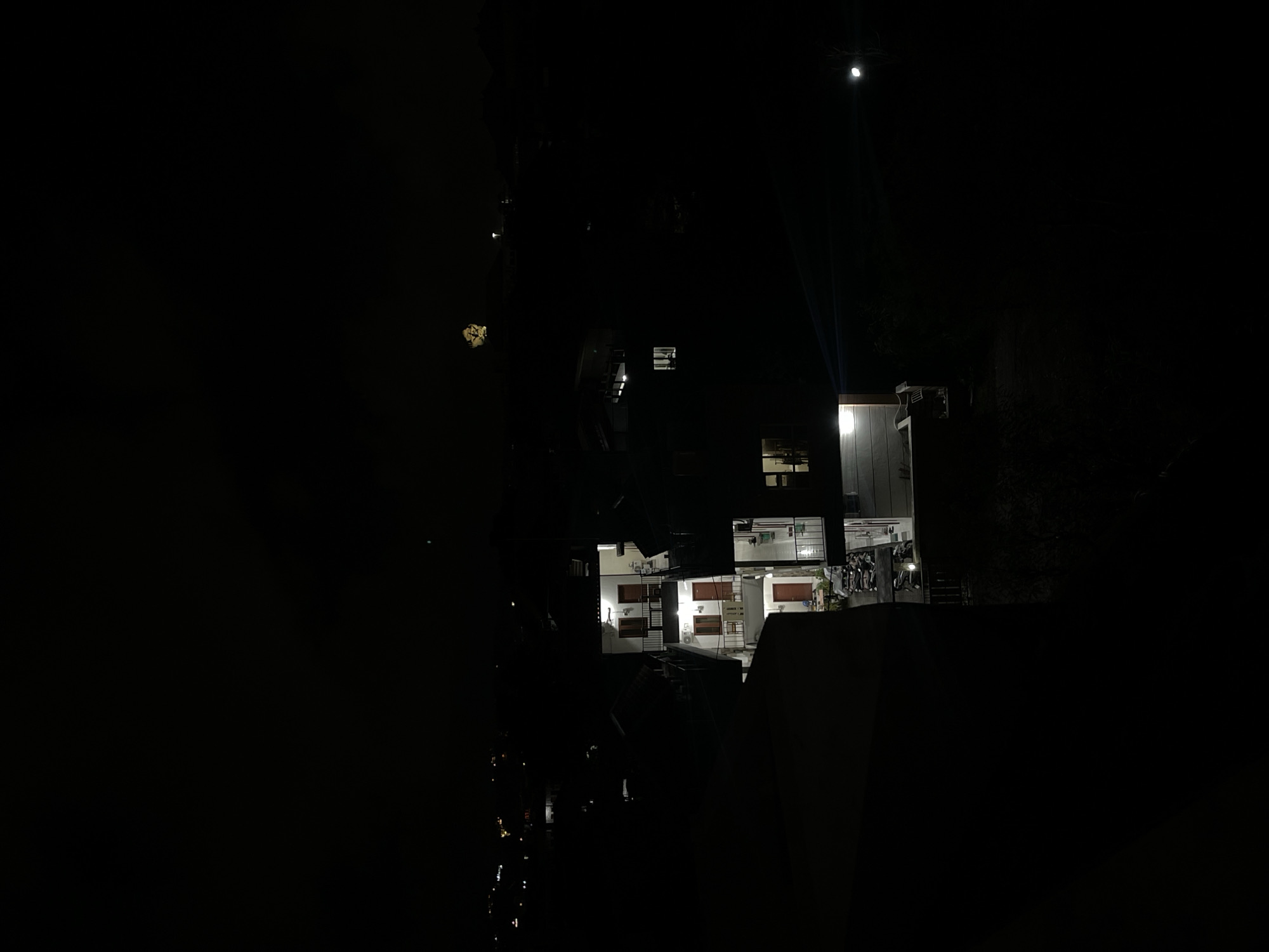 The image depicts a nighttime scene in the city of Kuta Selatan, Indonesia, characterized by darkness and minimal light sources. In the mid-ground, a multi-story building complex is visible, featuring illuminated windows and doorways, suggesting residential or commercial occupancy. The architecture appears to be typical of the region, with balconies and staircases connecting different levels. In the distance, faint lights of other buildings and streetlights are scattered across the landscape, indicating the urban setting. A notable element in the upper right portion of the image is what appears to be a distant fireworks display, indicated by a burst of yellow light against the dark sky. The foreground is dominated by dark, silhouetted structures, possibly part of adjacent buildings or balconies, obscuring a clear view of the immediate surroundings. The overall impression is one of a quiet night in a populated area, with a touch of celebratory ambiance from the distant fireworks.