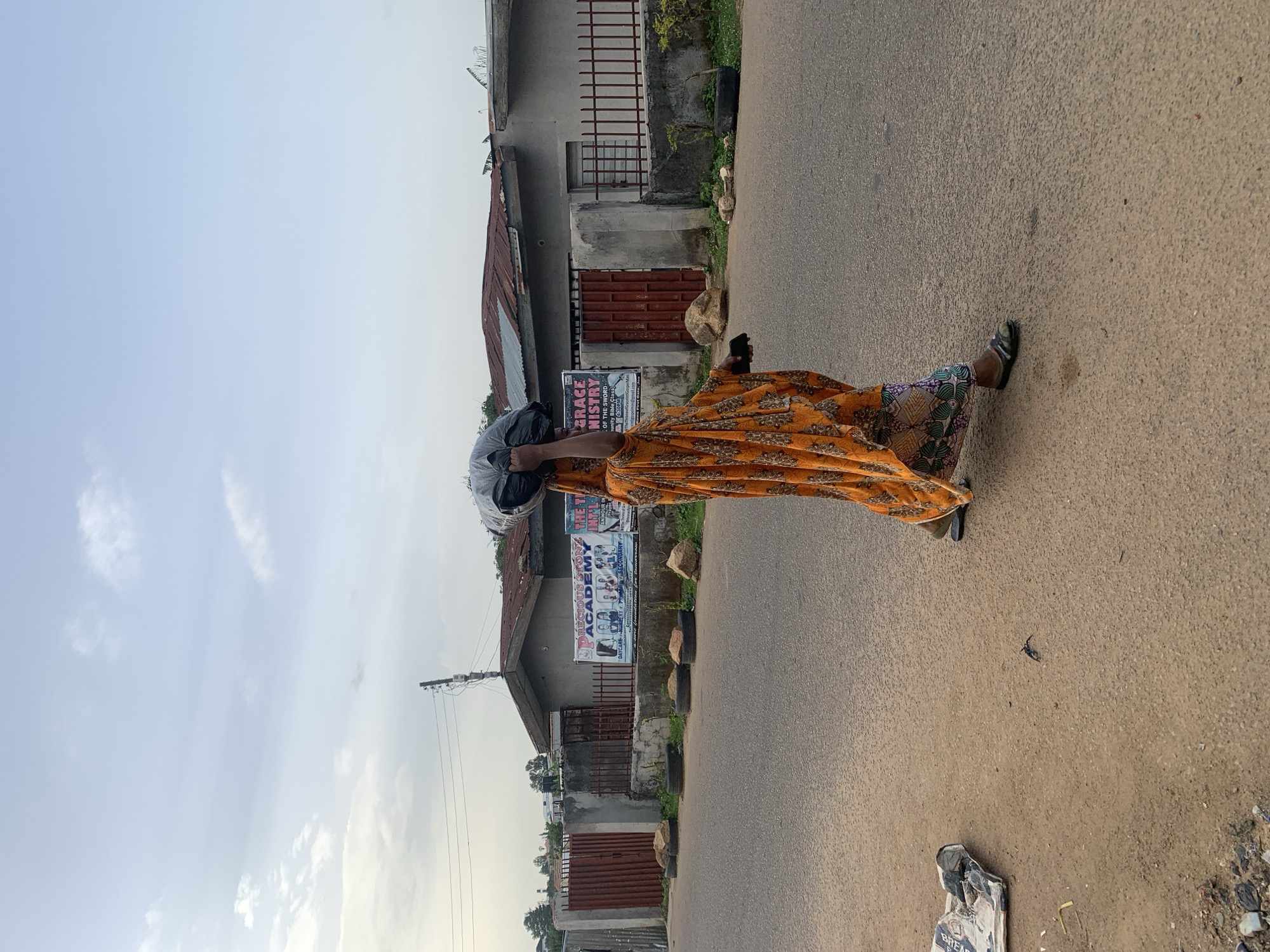 A woman in a yellow-orange printed dress and sandals walks on a paved road in Barakin Akawo, Nigeria. She carries a large, dark-colored bundle on her head. Behind her, there are buildings, including one with a sign that reads "PRECIOUS STONE ACADEMY". Utility poles with powerlines are visible against a cloudy sky. The road is cracked and shows signs of wear. Litter is visible on the roadside.