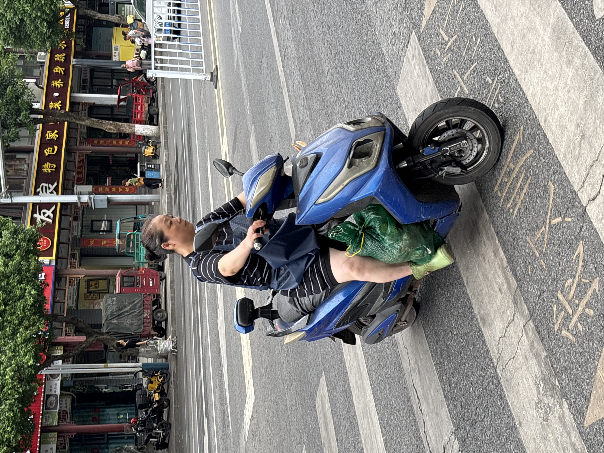 In the city of Jiangjin, China, a woman rides a blue electric scooter across a pedestrian crossing. She is wearing a black and white striped shirt, dark shorts, and bright green boots. A large green plastic bag is tied to the back of her scooter, and a dark blue bag hangs from the handlebars. The scooter has a blue and silver body. The street is paved with asphalt and marked with white pedestrian crossing stripes and some yellow painted symbols. In the background, there are buildings with awnings, trees, and other vehicles, including a red three-wheeled truck and a white car. The scene appears to be a typical urban street in China, with a mix of traditional architecture and modern elements.