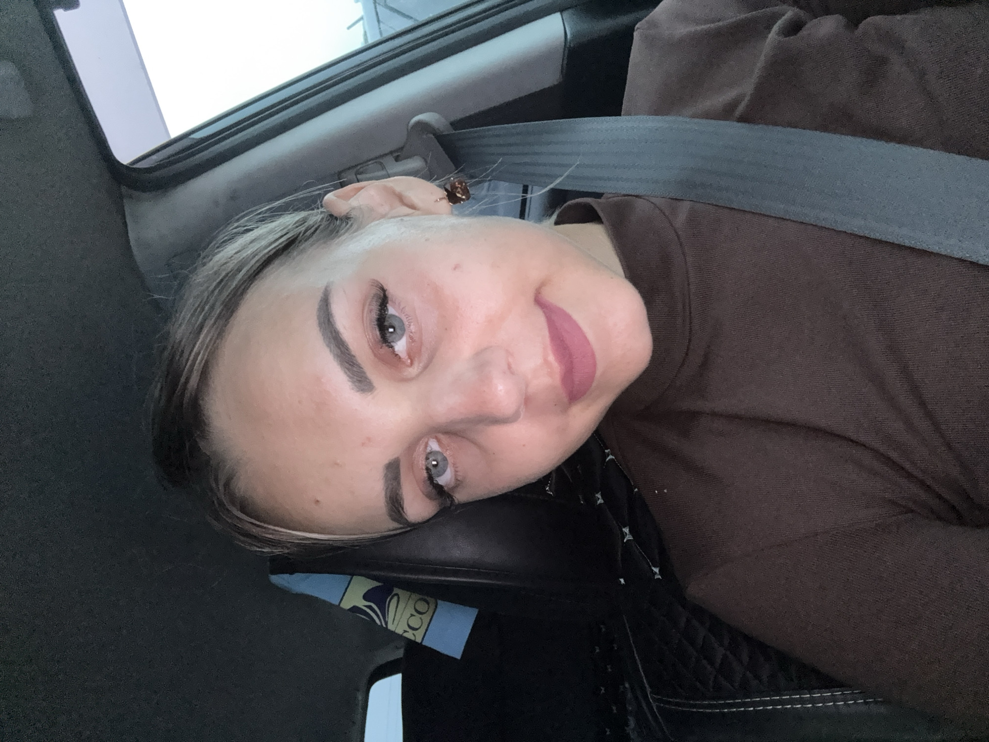 A woman is sitting in the driver's seat of a car, facing the camera. She has blonde hair pulled back in a bun, with a few strands framing her face. She is wearing a brown turtleneck shirt and a seatbelt is visible across her chest. Her makeup includes mascara, eyeliner, and a matte lipstick in a dusty rose shade. She has a small brown earring in her right ear. In the background, part of the car's interior is visible, including a black leather seat with a quilted pattern and a blue and yellow fabric item with the word "ECO" printed on it. The lighting suggests it is daytime, possibly overcast, and the background outside the car is out of focus and appears to be a bright, neutral sky. The image appears to be a selfie taken inside a vehicle, likely in a parking lot or on a road. The location is Yuzhno-Sakhalinsk, Russia.