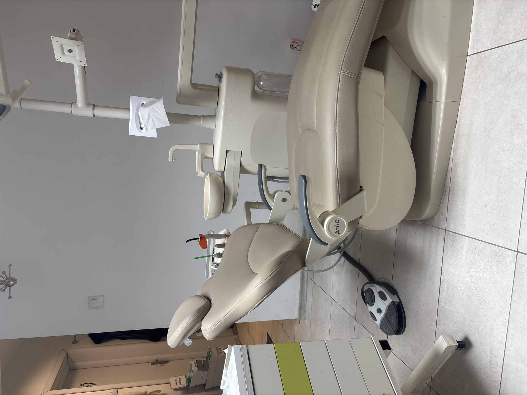 The image shows a dental examination room, most likely in a clinic or office in Sofia, Bulgaria. The primary focus is a beige-colored dental chair with attached equipment. A paper towel dispenser is visible on the equipment arm. The dental chair has a foot pedal controller on the tiled floor. A storage cabinet with visible supplies is in the foreground. The walls are painted in a light color. Overall, the room appears clean and functional.