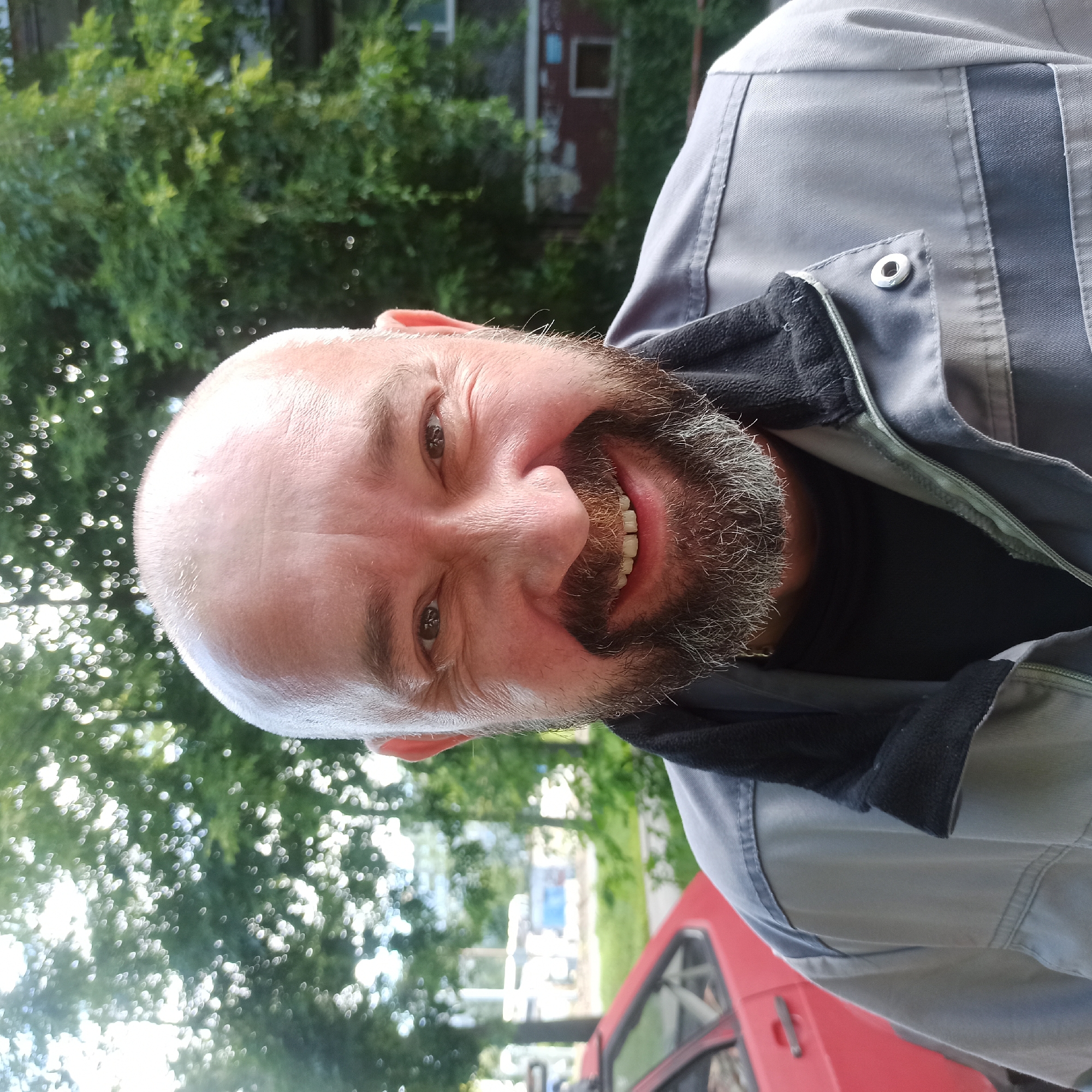 A middle-aged bald Caucasian man with a full beard is pictured outdoors, close to a red car, with trees and a building in the background. He is wearing a grey work jacket with a black collar and a dark t-shirt underneath. The man is smiling directly at the camera, with his eyes crinkled at the corners. The background suggests an urban setting, possibly in Nizhny Novgorod, Russia, with dappled sunlight filtering through the leaves of the trees. The red car appears to be parked, and the building in the background is a multi-story structure with visible windows. The overall impression is of a candid outdoor portrait.