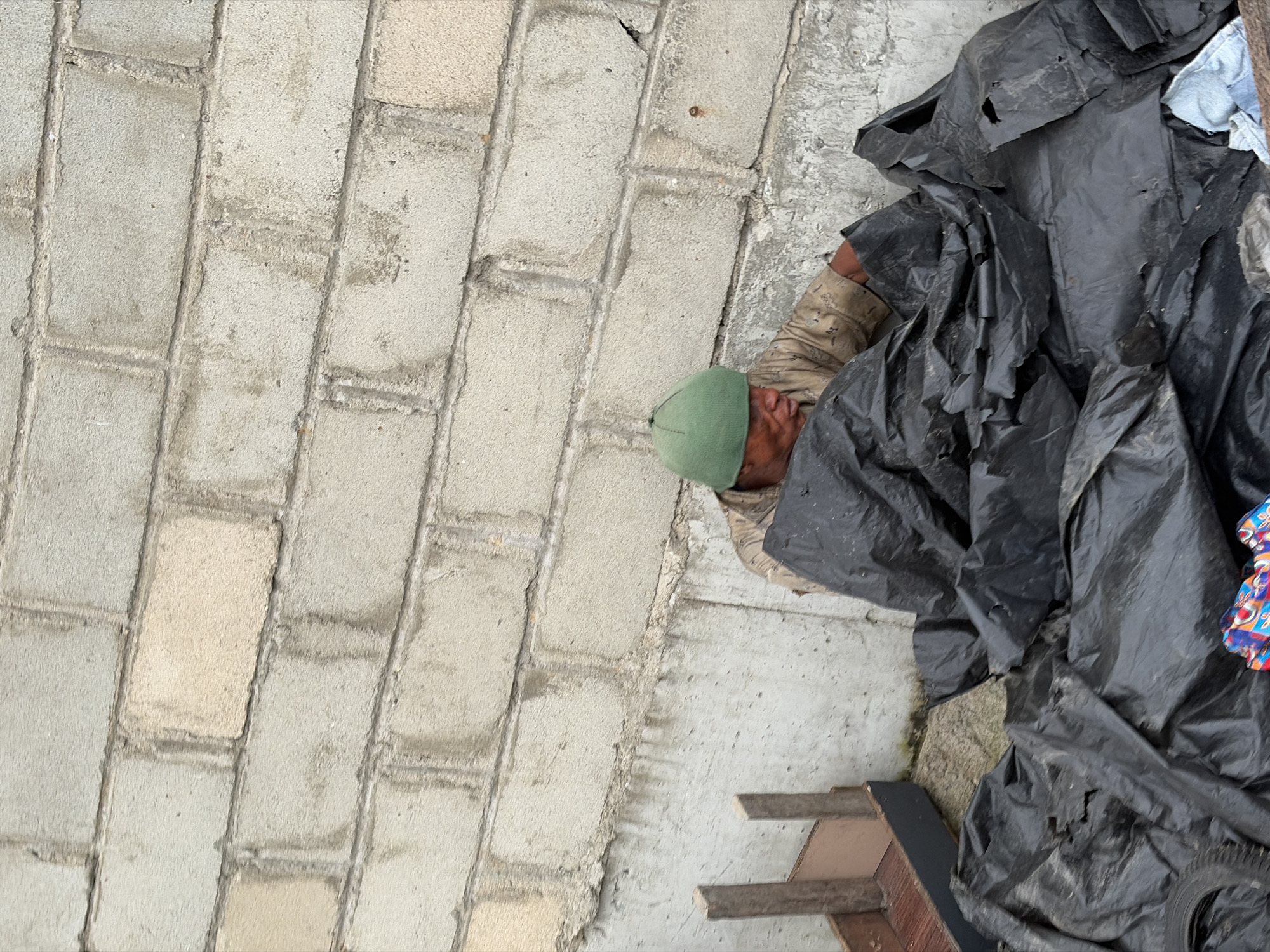 A person wearing a green beanie and a light brown, patterned shirt is partially obscured by black plastic sheeting. They are kneeling or sitting on the ground, with their head and upper body visible. The person appears to be interacting with the plastic, possibly gathering or arranging it. The background consists of a rough, grey concrete block wall with visible mortar lines and some staining. To the left of the person, a wooden structure with dark grey and brown components is visible. The ground appears to be a mix of dirt and debris, with some of the black plastic sheets covering a large portion of the foreground, including what looks like a tire on the left. The scene is likely outdoors, given the wall and the natural ground surface. The location is identified as Igboefon, Nigeria.