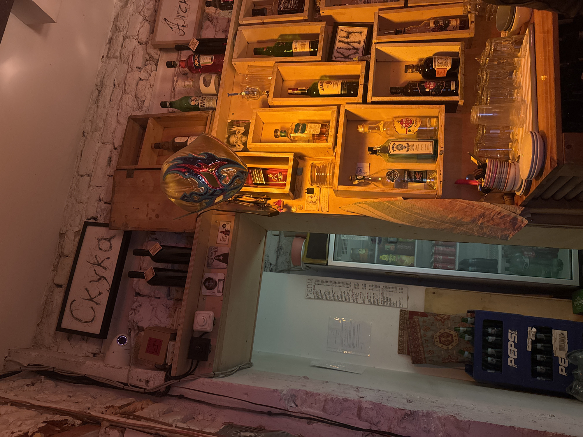 Here's a detailed description of the image:
**Location and Environment:**
The photo appears to be taken inside a bar or restaurant, potentially located in Sofia, Bulgaria, judging by the Cyrillic script visible in some areas. The environment has a slightly aged or rustic aesthetic, evident in the exposed brick walls and weathered wooden shelving. The lighting is dim, creating an intimate atmosphere.
**Objects:**
* **Shelving:** The primary element is a series of wooden shelves or boxes mounted on the wall, which serve as storage and display for various items.
* **Liquor and Drinks:** A variety of liquor bottles (whiskey, rum, etc) are arranged on the shelves. There are glass bottles of what appears to be beer stored inside the refrigerator. A blue Pepsi crate is visible below the serving window.
* **Glassware:** A collection of drinking glasses, cups, and saucers are stacked near the right edge of the shelves.
* **Menu:** There is a handwritten or printed menu visible through a small opening, likely leading to the bar area.
* **Text and Decorations:** Cyrillic text is noticeable in several places, including a framed piece on the left wall with the word "Скука" and a plaque on the top shelf with "Алчн."
* **Other Items:** A decorative mask, electrical sockets, light switch, and CCTV security camera.
**Summary**
The image portrays a dimly lit bar or restaurant interior, likely in Sofia, Bulgaria, with liquor bottles, glassware, and decorative items arranged on wooden shelving. The Cyrillic text and architectural details provide clues to the location.