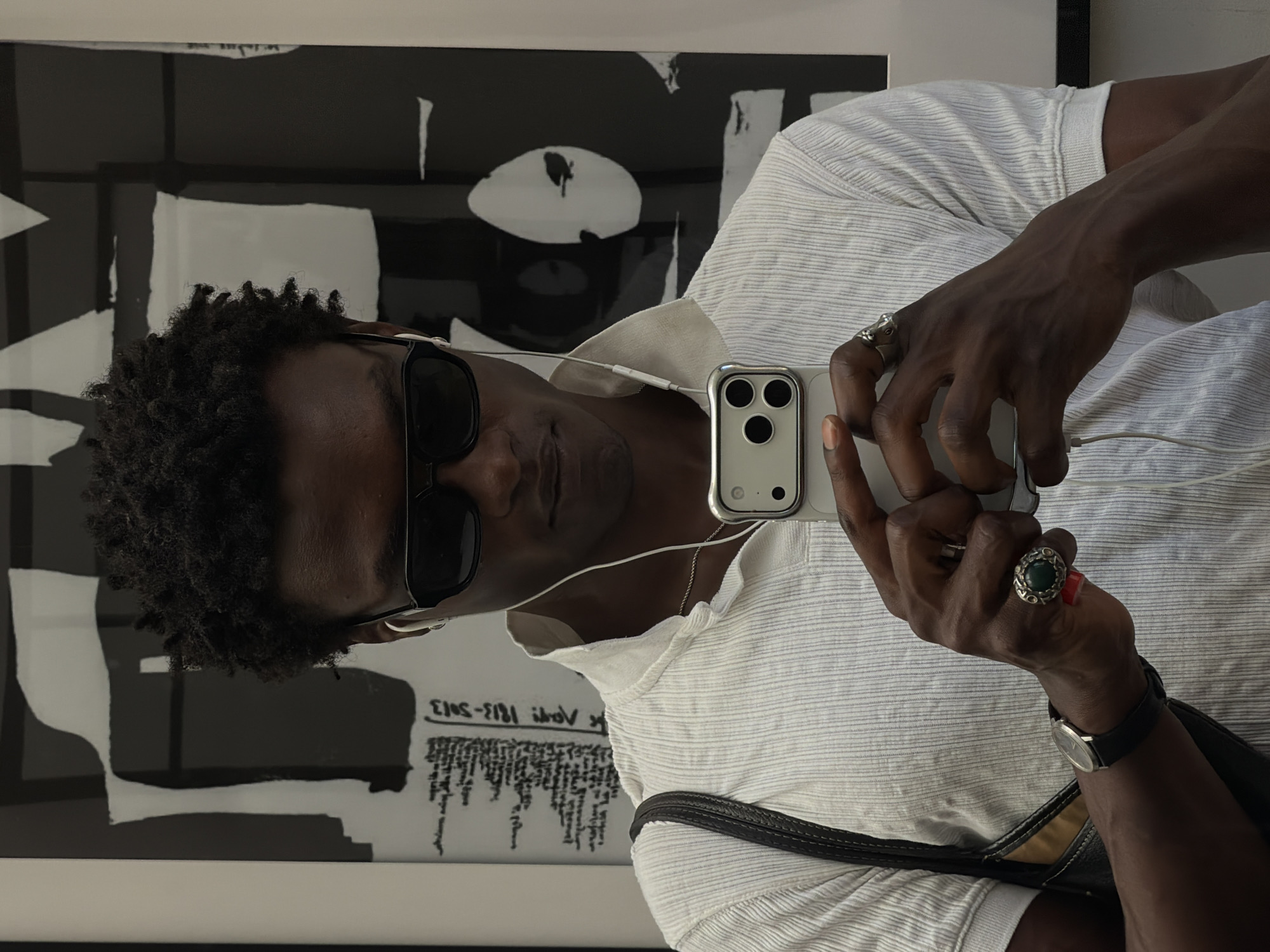 This is a vertical, medium close-up shot of a Black person holding a smartphone and taking a selfie. The person is wearing sunglasses and a light-colored, collared shirt with a textured pattern. They have short, curly dark hair and are wearing two rings on their right hand, one with a green stone and the other a silver band. A watch with a black band is also visible on their wrist. The phone's camera is visible, as are white earbuds connected to it.
The background features a large piece of abstract art with bold black and white shapes. Some text is visible on a white surface in the background, partially obscured by the person's arm and phone. The legible text appears to be handwritten or printed in a small font, with some characters visible including "2013" and "5013" as well as some illegible cursive.
The lighting suggests it is daytime, possibly indoors given the clear focus and lack of harsh shadows. The overall impression is casual, possibly a moment captured while exploring an art exhibition or a similar cultural venue. The person’s expression is neutral, as they are focused on taking the photo. The setting appears to be a gallery or exhibition space in London, United Kingdom.