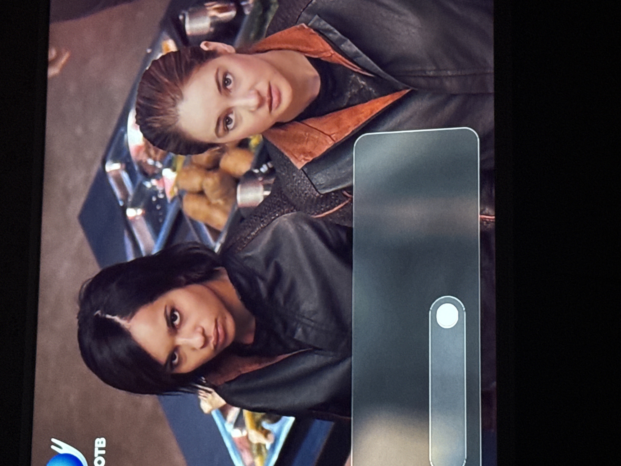 Two women are standing next to each other, wearing leather jackets with orange lining. There is a tray of food between them. A gray, rectangular object with a white button is in the right foreground. "OTB" is visible at the bottom left. The location is Mytishchi, Russia.