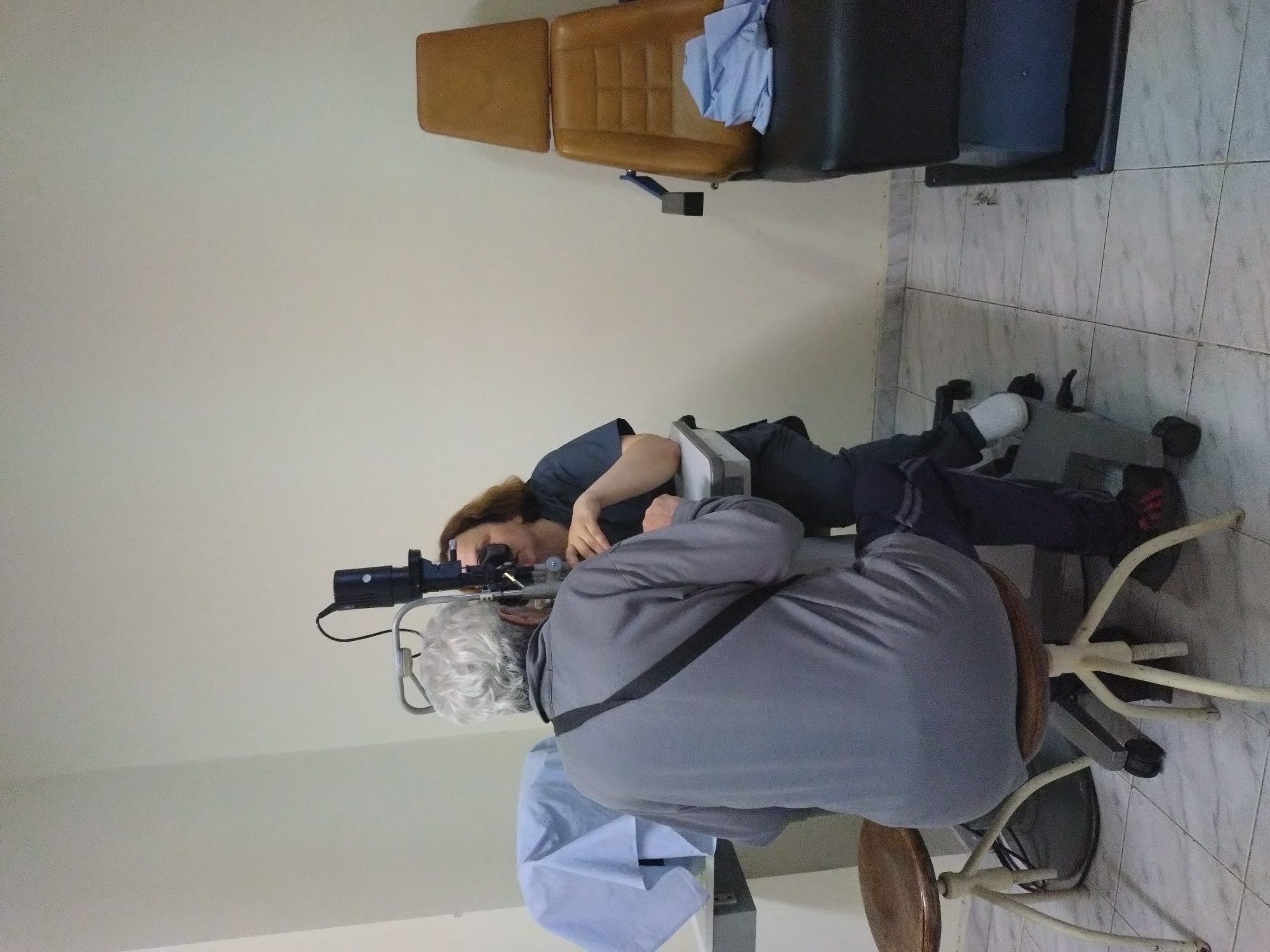 A person is lying on a table with a headrest in a medical office. Another individual is seated behind the patient, using a medical device to examine the patient's eyes. The office contains a chair and medical equipment. The image does not provide enough details to determine the location in Stara Zagora, Bulgaria.