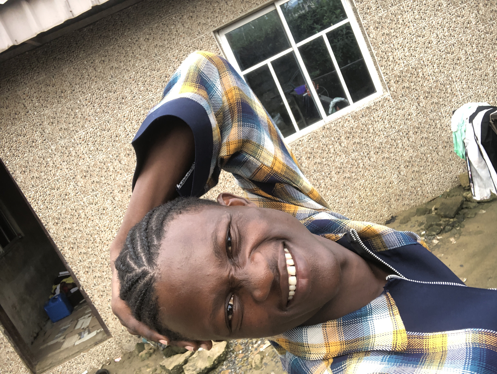 A young Black male is pictured from the chest up, outdoors, in what appears to be Ado, Nigeria. He is facing the camera and smiling widely, with his hand resting on the back of his head. He has dark skin, braided hair, and is wearing a blue, yellow, and white plaid shirt over a navy blue zipper vest. Behind him, the textured wall of a building is visible, with a white-framed window that has several panes. The window offers a view of green foliage. In the upper left corner, a deep alcove in the wall contains a blue plastic container and some other items. A small patch of muddy ground is visible in the lower left quadrant of the image.