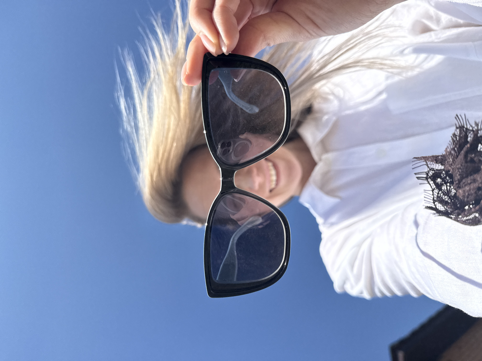 A low-angle, close-up shot shows a woman holding up sunglasses against a clear blue sky, likely in the city of Chaikovsky, Russia. The woman is slightly out of focus behind the sunglasses but visible enough to discern she is smiling. She wears a white blouse with a dark lace detail on the side. Her blonde hair is partially blurred due to movement. The sunglasses are large and dark-framed. The reflection in the lens reveals a cell phone camera. A small section of a dark building roof is visible in the extreme bottom of the frame.
