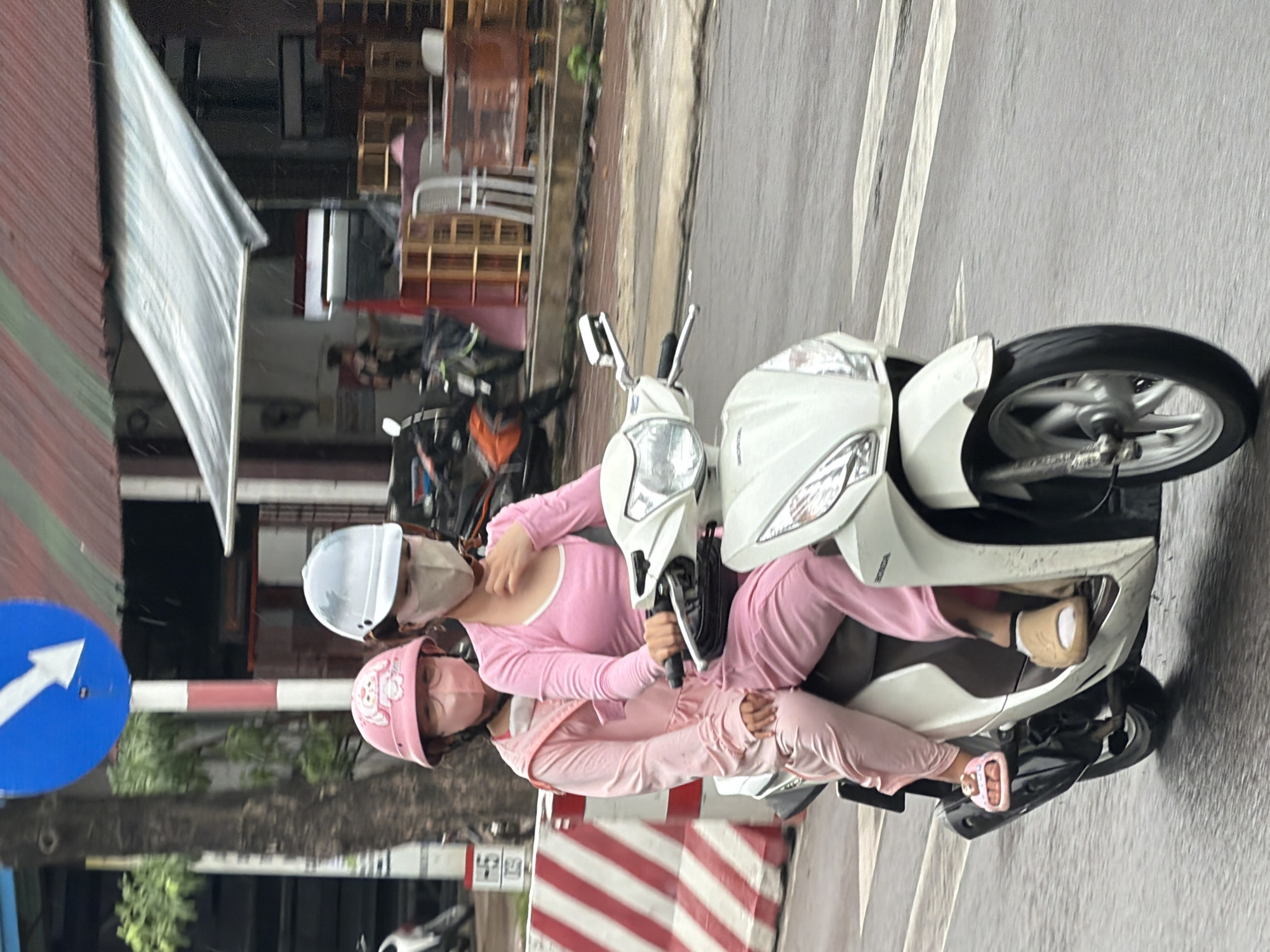The image depicts two individuals on a white Honda scooter on a wet street in Nha Trang, Vietnam. The person in the front, likely the driver, wears a light-colored face mask, a silver helmet, and a pink long-sleeved shirt. The person behind, a child, wears a pink helmet with a cartoon character on it, a pink face mask, and a pink jumpsuit. Both individuals are wearing masks covering their faces. The scooter has a white body with black accents and is moving forward on a wet road, indicated by reflections on the surface. The background shows a building with a corrugated metal roof, some wooden shelves with items, and a traffic sign with a white arrow pointing down and to the left on a blue circular background. There are also some red and white striped bollards along the side of the road. The overall lighting suggests it is daytime, possibly overcast or raining lightly.