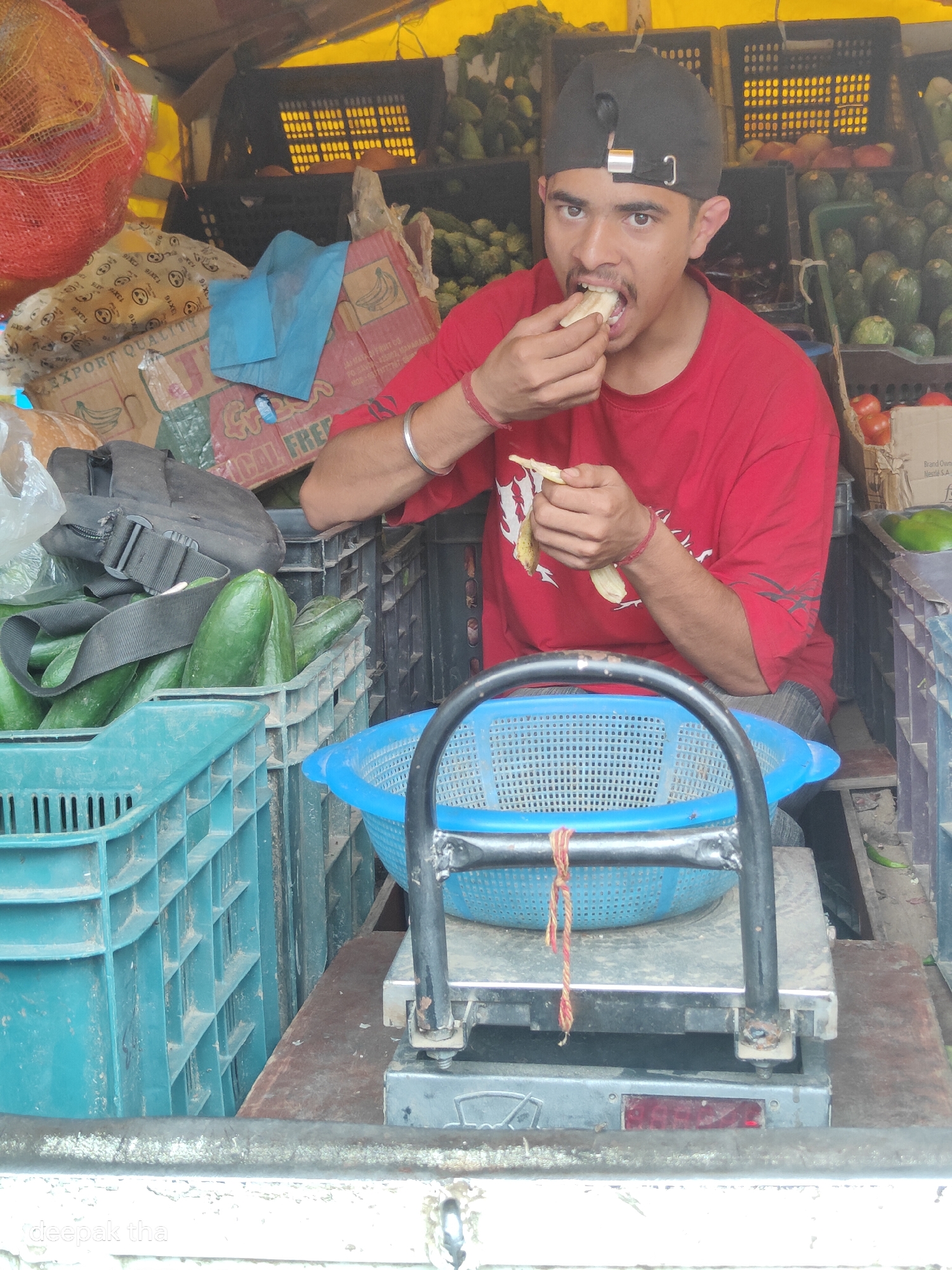 A young man with a black baseball cap, wearing a red t-shirt, is seated in what appears to be a market stall in Vashisht, India. He is in the process of eating a piece of peeled banana, holding it in his right hand near his mouth and a second piece in his left hand. The stall is filled with crates of produce, including large green cucumbers in a teal crate to his left, and other vegetables and fruits are visible in the background. A blue plastic basket is positioned on a scale in front of him. The overall impression is of a bustling, informal marketplace setting.